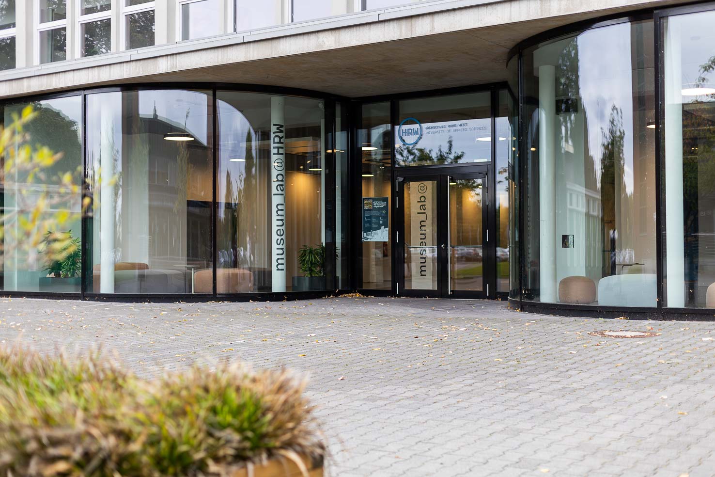 Eingangsbereich des Campus Mülheim (Parkstadt) der Hochschule Ruhr West: moderne Glasfassade mit geschwungener Front, Doppeltür und Beschriftung ‚museum.lab @ HRW‘.