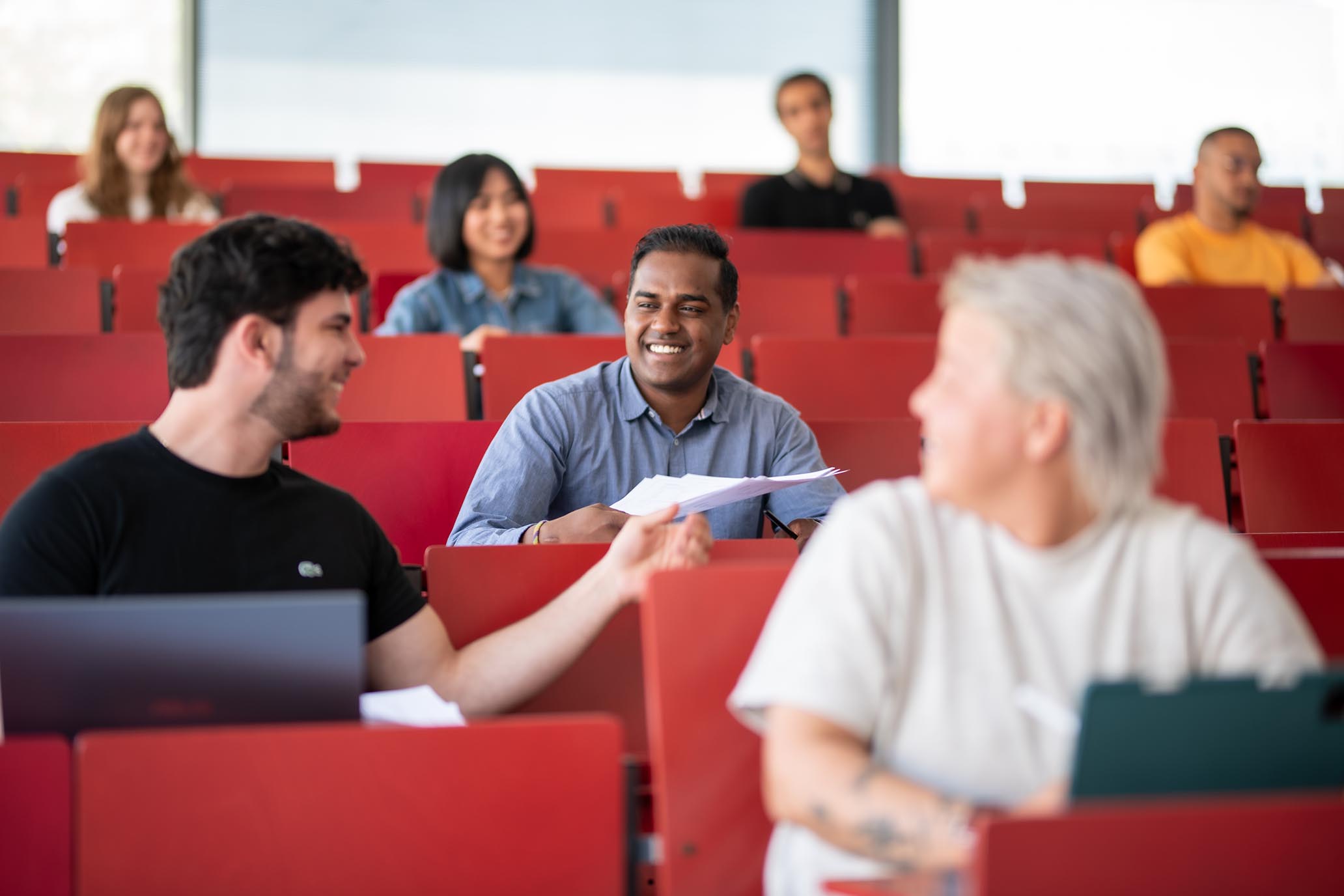 Mehrere Personen sitzen in einem Hörsaal. Eine Person gibt ein Blatt weiter mach hinten.