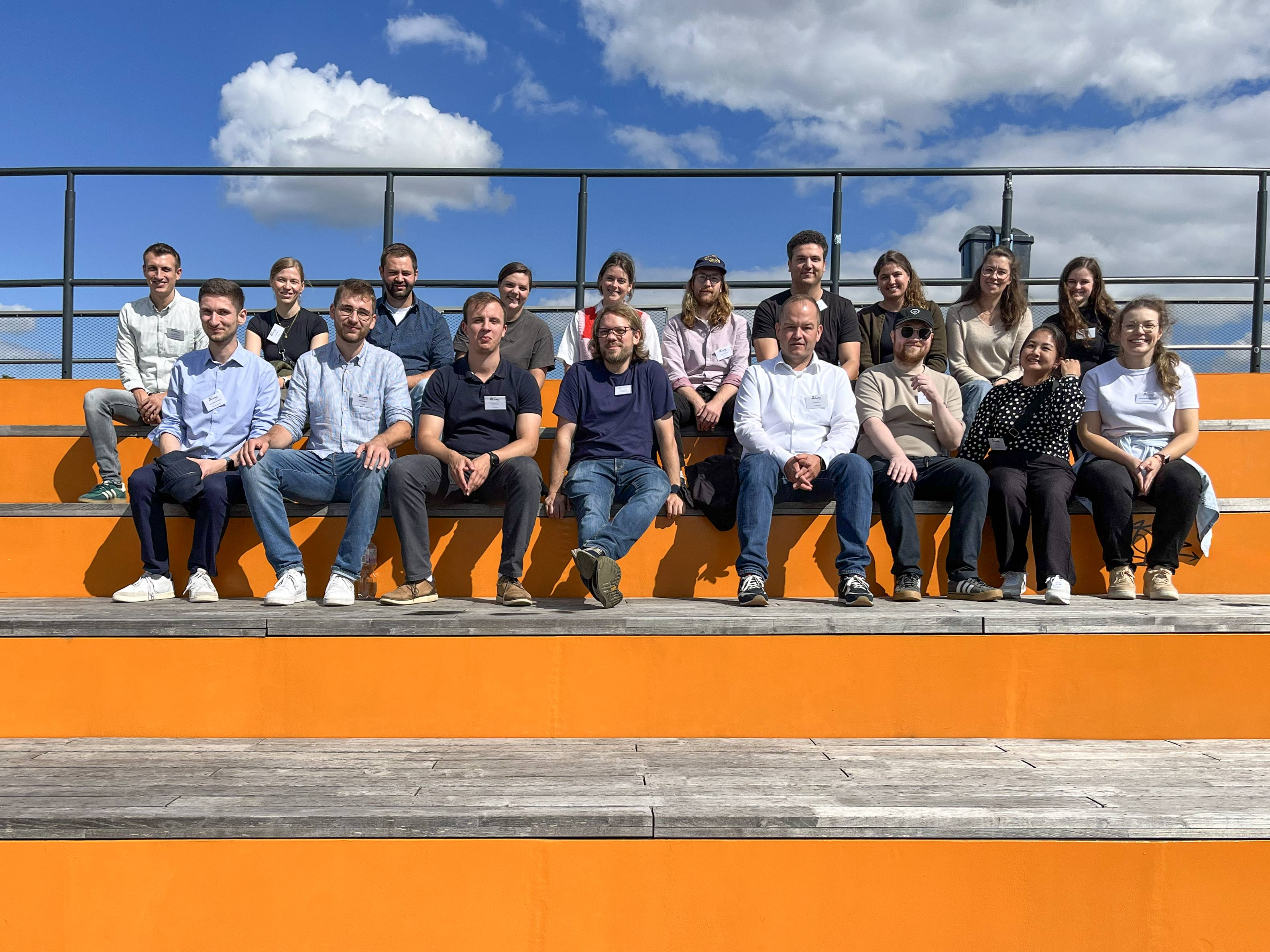 Gruppenfoto: Menschen sitzen auf orangefarbener Tribüne im Freien unter blauem Himmel mit Wolken.