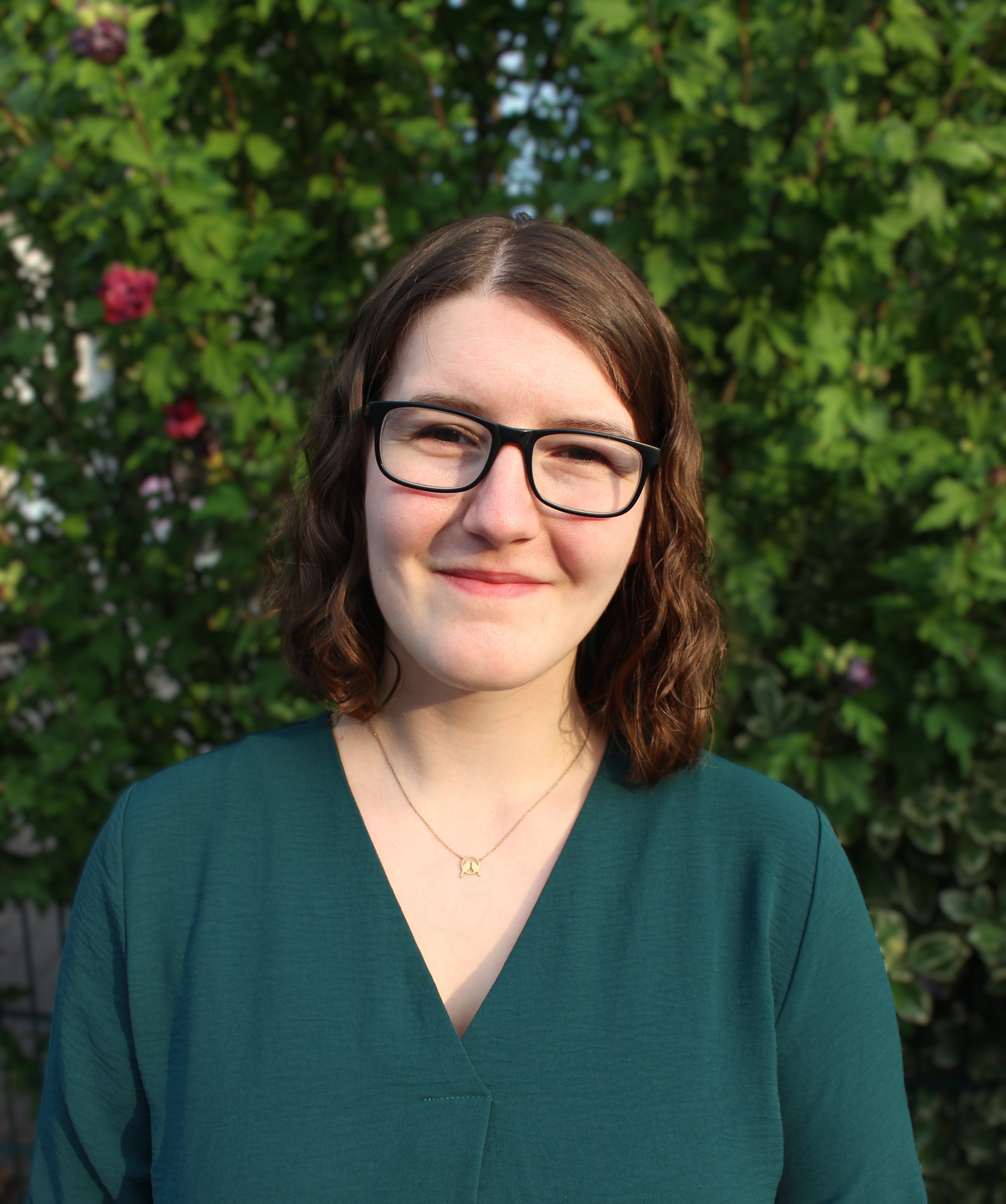 Porträt einer lächelnden Person mit Brille, schulterlangen Locken und Kette im grünen Oberteil vor dichtem Laub im Freien.
