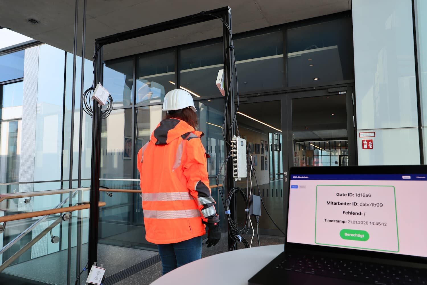 Person mit Helm und Warnjacke durchläuft Zugangsschleuse am Gebäude; im Vordergrund Laptop mit Zutrittskontrolle, Status: Berechtigt.