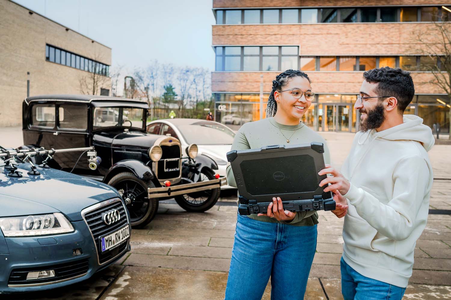 Eine Studentin hält einen Laptop, während sie mit einem anderen Studenten vor drei Autos auf dem Campus Mülheim steht.