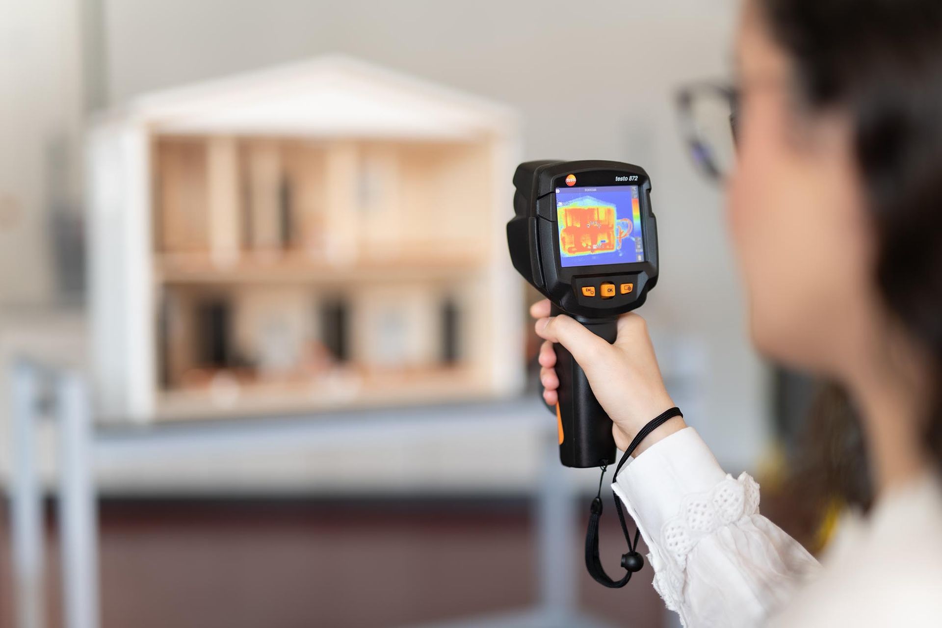 Eine Studentin der Hochschule Ruhr West nutzt eine Wärmebildkamera zur Analyse eines Modellhauses. Das Bild zeigt praxisnahes Lernen im Bereich Energieeffizienz und Gebäudetechnik an der HRW.