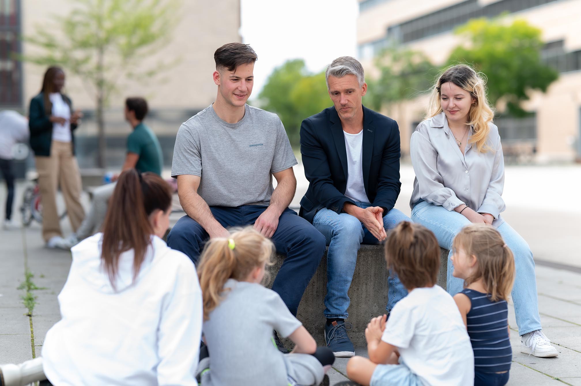Ein Mann sitzt mit Studenten und spricht mit Kindern auf dem Campus Mülheim.