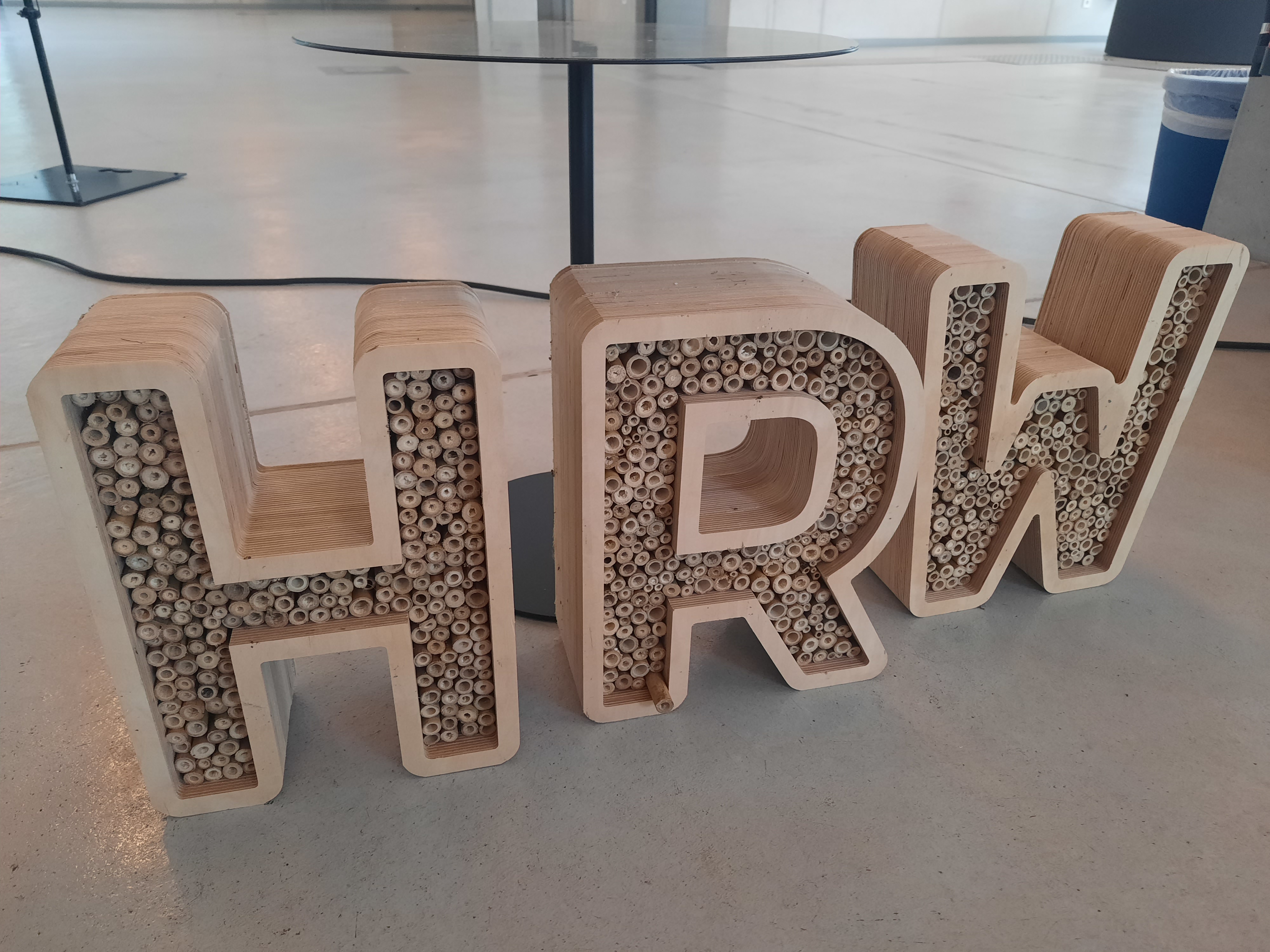 Holzbuchstaben „HRW“ als Insektenhotel gestaltet, gefüllt mit zahlreichen Bambusröhrchen. Die Buchstaben stehen auf einem hellen Hallenboden vor einem runden Tisch.
