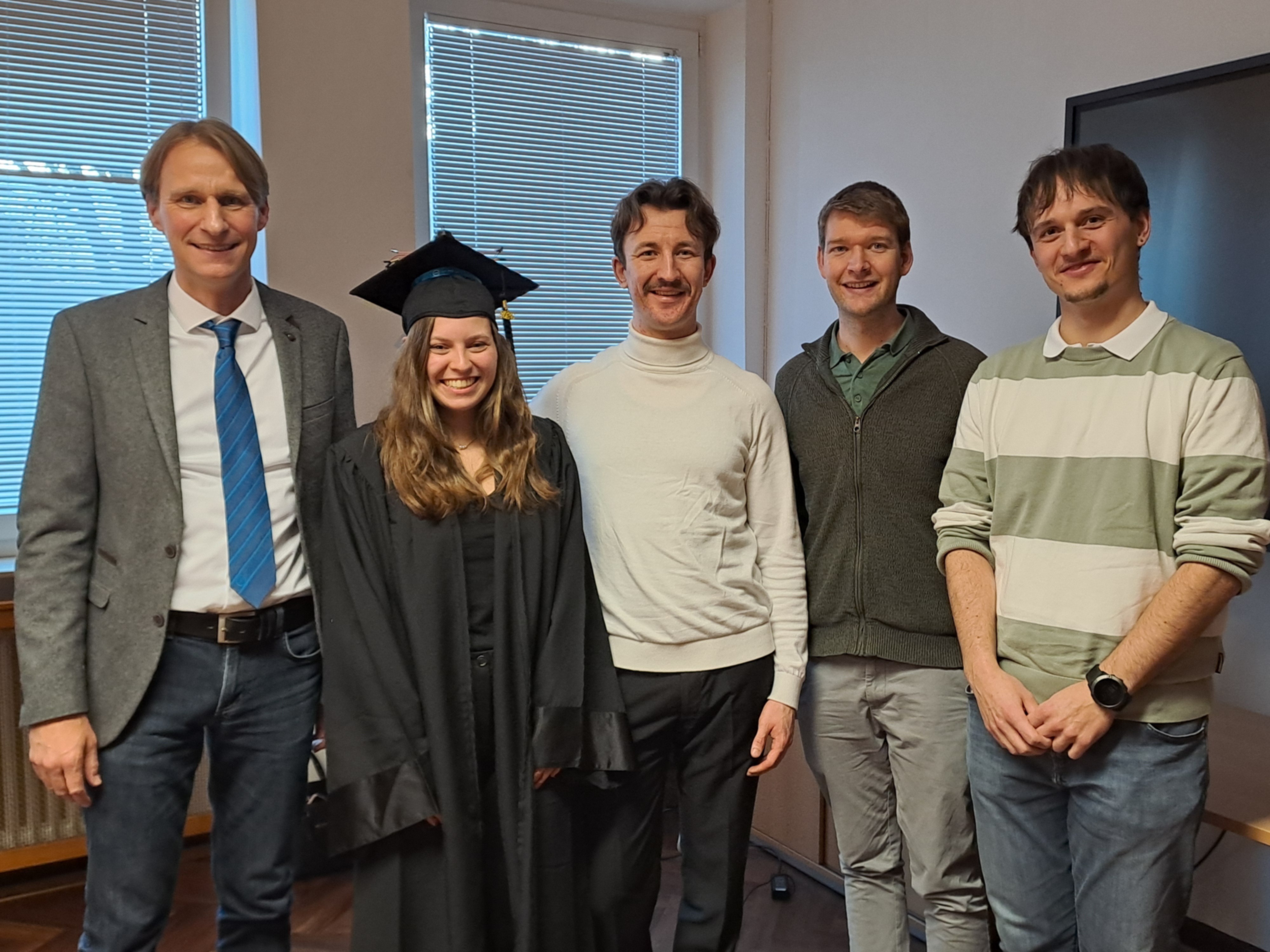 Eine Frau mit Doktorhut steht gemeinsam mit vier Männern in einem Unterrichtsraum.