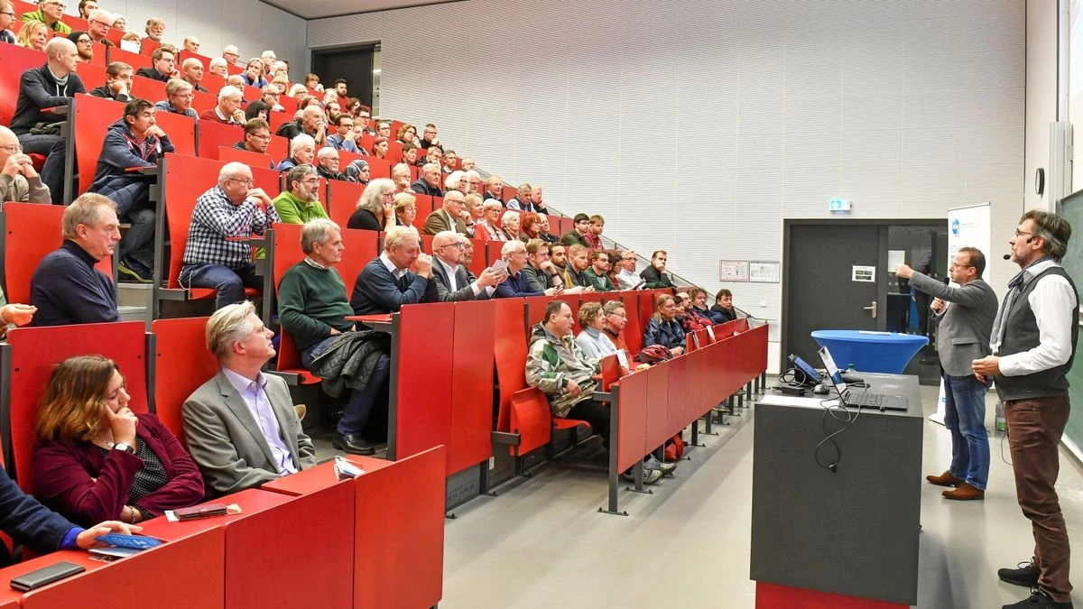 Gut besetzter Hörsaal mit roten Sitzreihen; zwei Vortragende vorn, Publikum hört zu, einige heben die Hand und diskutieren.