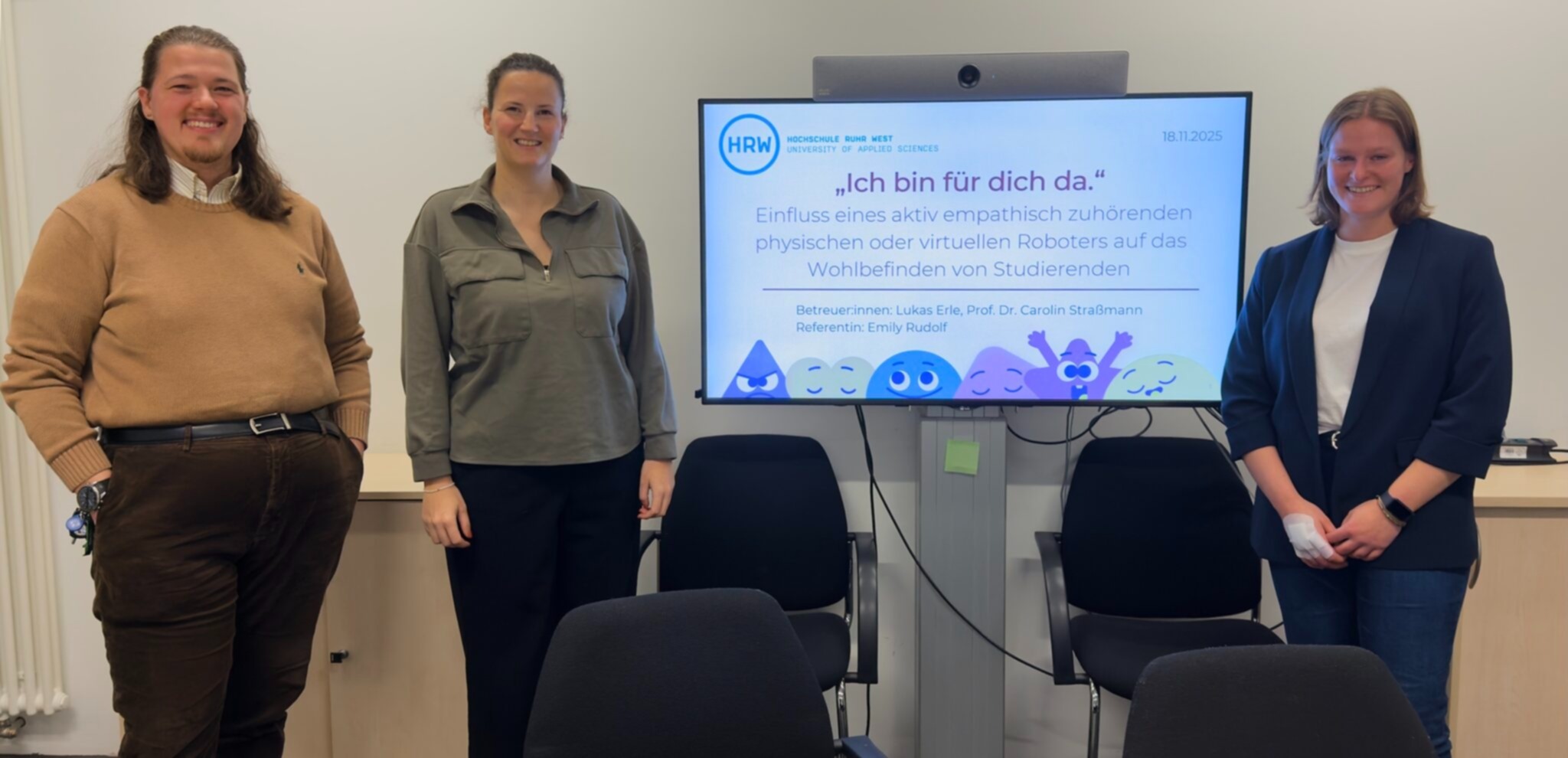 Drei Personen stehen lächelnd in einem Seminarraum vor einem großen Monitor. Auf dem Bildschirm ist die Titelseite einer wissenschaftlichen Präsentation der Hochschule Ruhr West zu sehen, die sich mit dem Einfluss empathischer physischer oder virtueller Roboter auf das Wohlbefinden von Studierenden beschäftigt. Auf dem Monitor ist zu lesen "Ich bin für dich da."