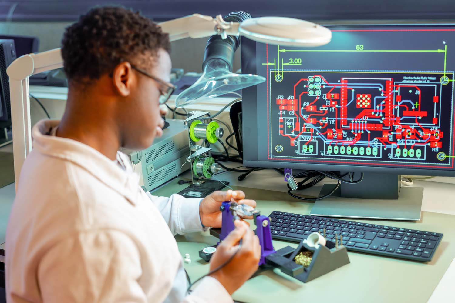 Ein Student arbeitet an einer elektronischen Platine, und vor ihm ist die Darstellung der Platine auf einem Computerbildschirm zu sehen.