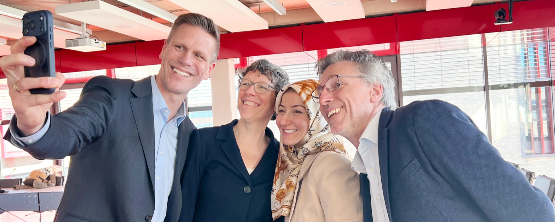 Zwei Männer und zwei Frauen schauen in eine Handykamera, ein Mann macht ein Selfie von allen Personen