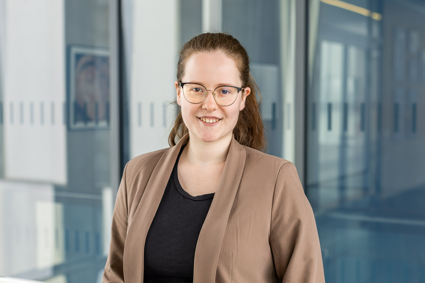 Porträtfoto einer lächelnden Person mit Brille im braunen Blazer vor Glaswänden in einem modernen Büro.
