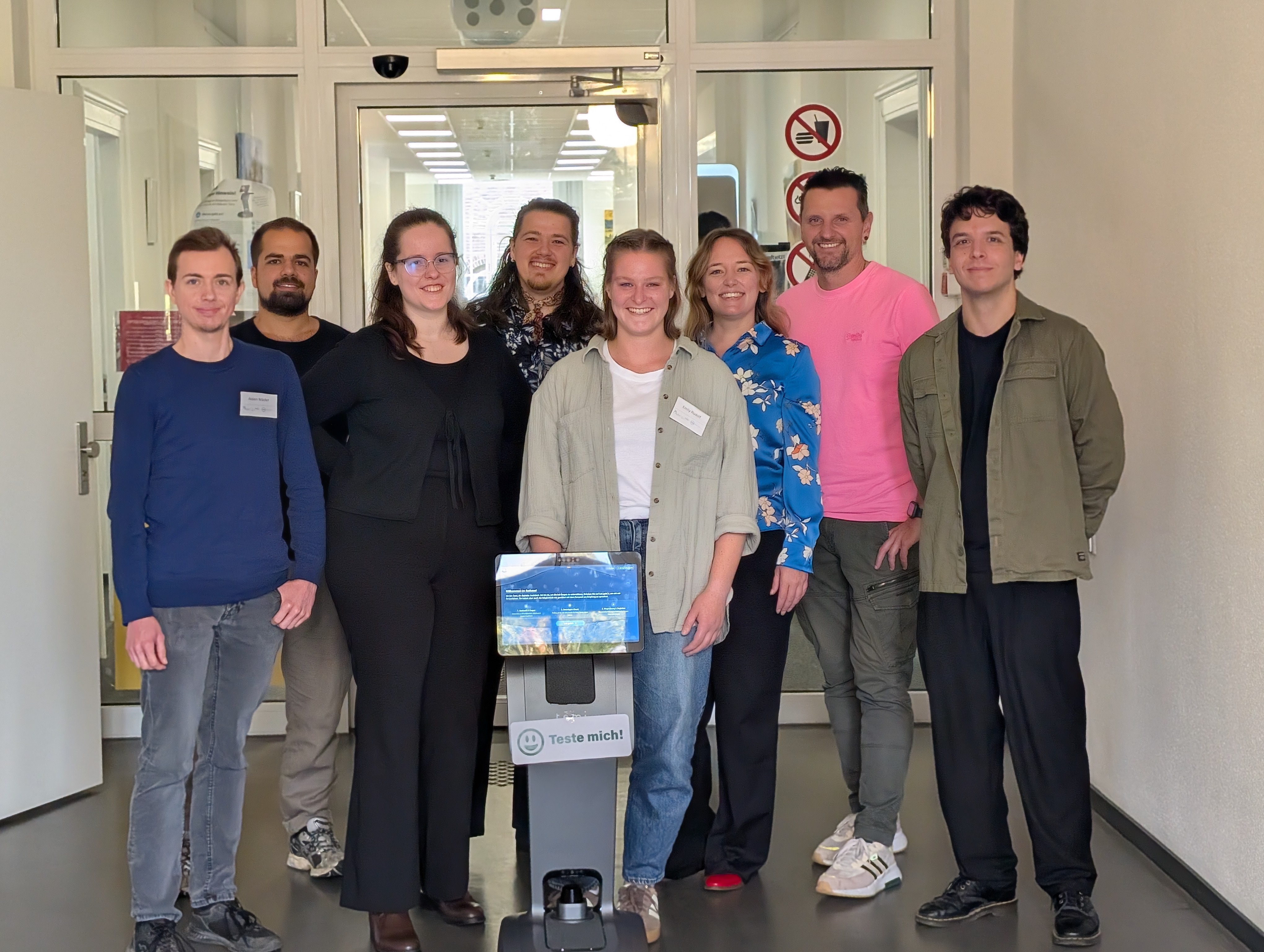Studierendengruppe stehen zusammen mit Roboter Temi und den betreuenden Lehrenden im Flur der Stadtverwaltung Bottrop