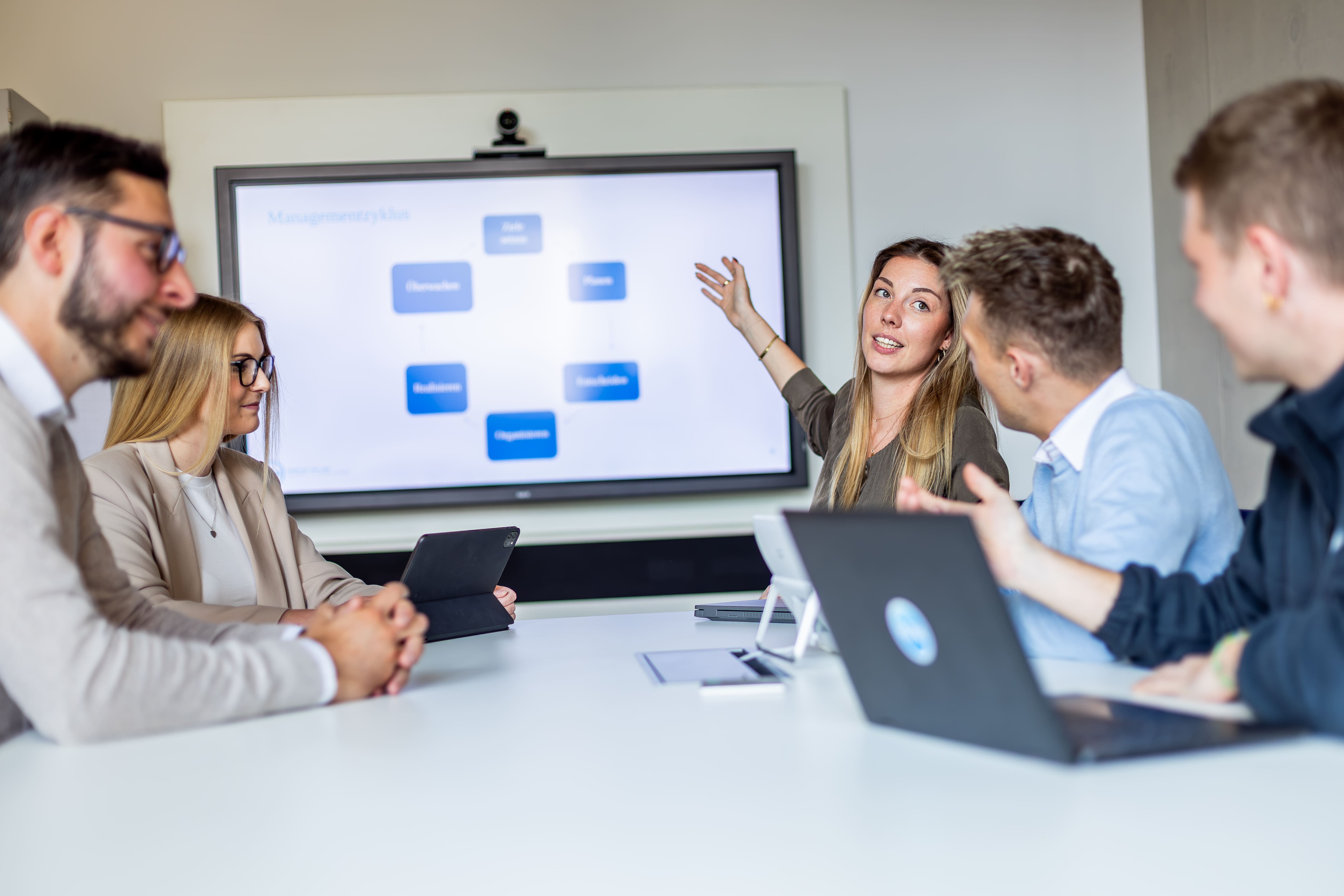 Eine junge Frau präsentiert in einem Besprechungsraum vor einer kleinen Gruppe. Sie zeigt auf eine Folie mit einem Managementzyklus auf einem Bildschirm, während vier Kolleginnen und Kollegen aufmerksam zuhören. Auf dem Tisch liegen Laptops und Tablets.