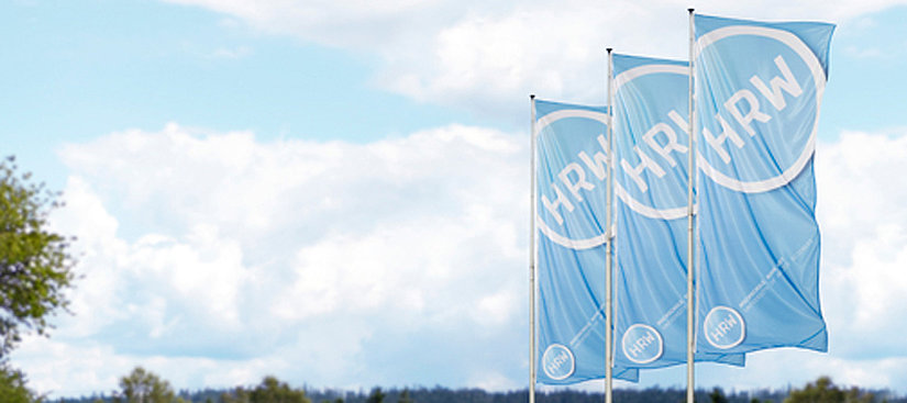 Auf dem Bild ist eine schöner blauer Himmel zusehen und rechts im Vordergrund befinden sich drei große hellblaue HRW Flaggen.