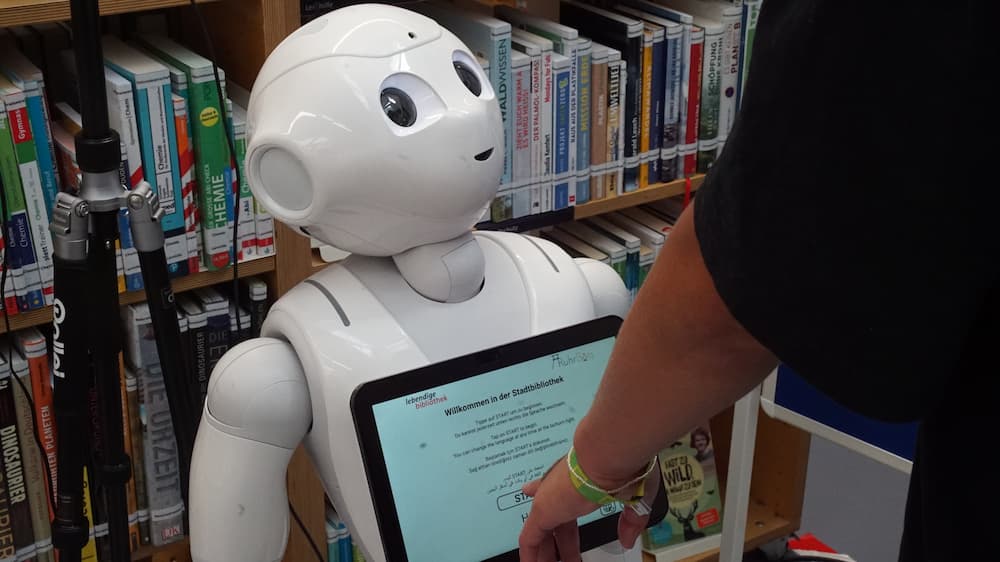 Ein weißer Roboter steht vor einem Bücherregal. In Brusthöhe ist ein Bildschirm angebracht auf dem Bibliotheksinhalte zu sehen sind. Eine Person, von der nur der Arm und die Hand zu sehen ist, drückt auf den Touchscreen.