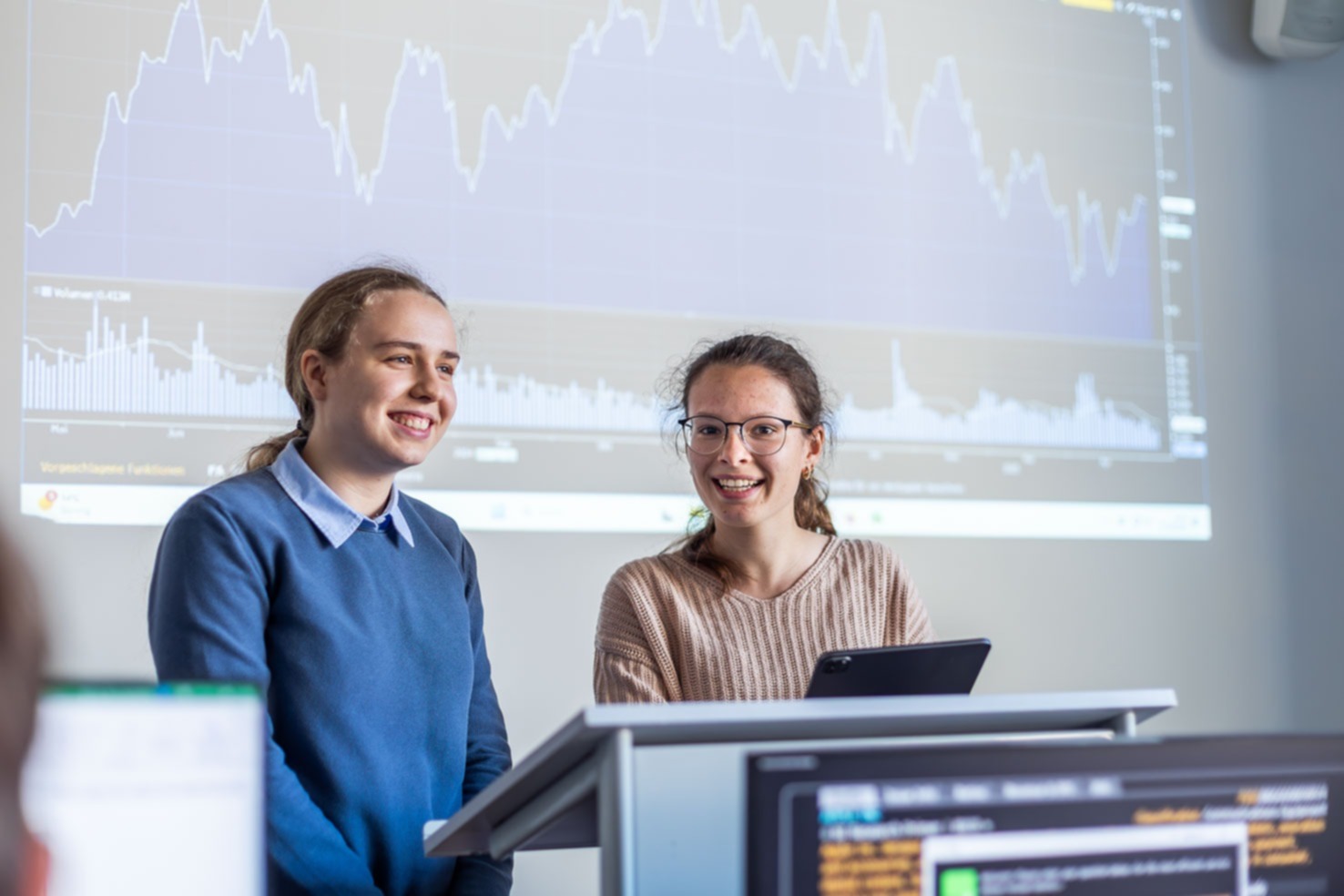 Zwei Frauen präsentieren und diskutieren Analyseergebnisse vor einer projizierten Finanzgrafik; im Vordergrund stehen Monitore mit Auswertungen, die Szene zeigt datenbasiertes Arbeiten im Kontext Finanzcontrolling.