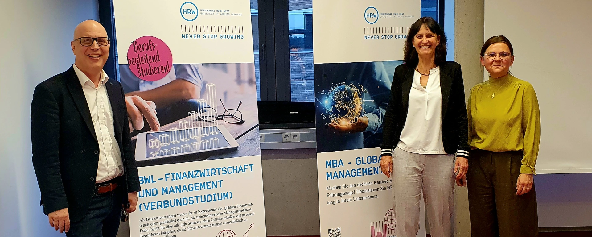Drei Personen stehen in einem Innenraum vor zwei Roll-up-Bannern der Hochschule Ruhr West. Links steht ein Mann im dunklen Anzug und mit Brille, rechts daneben zwei Frauen. Auf den Bannern sind Studienangebote der HRW zu sehen, darunter „BWL – Finanzwirtschaft und Management“ sowie „MBA – Global Management“. Alle drei blicken in die Kamera.