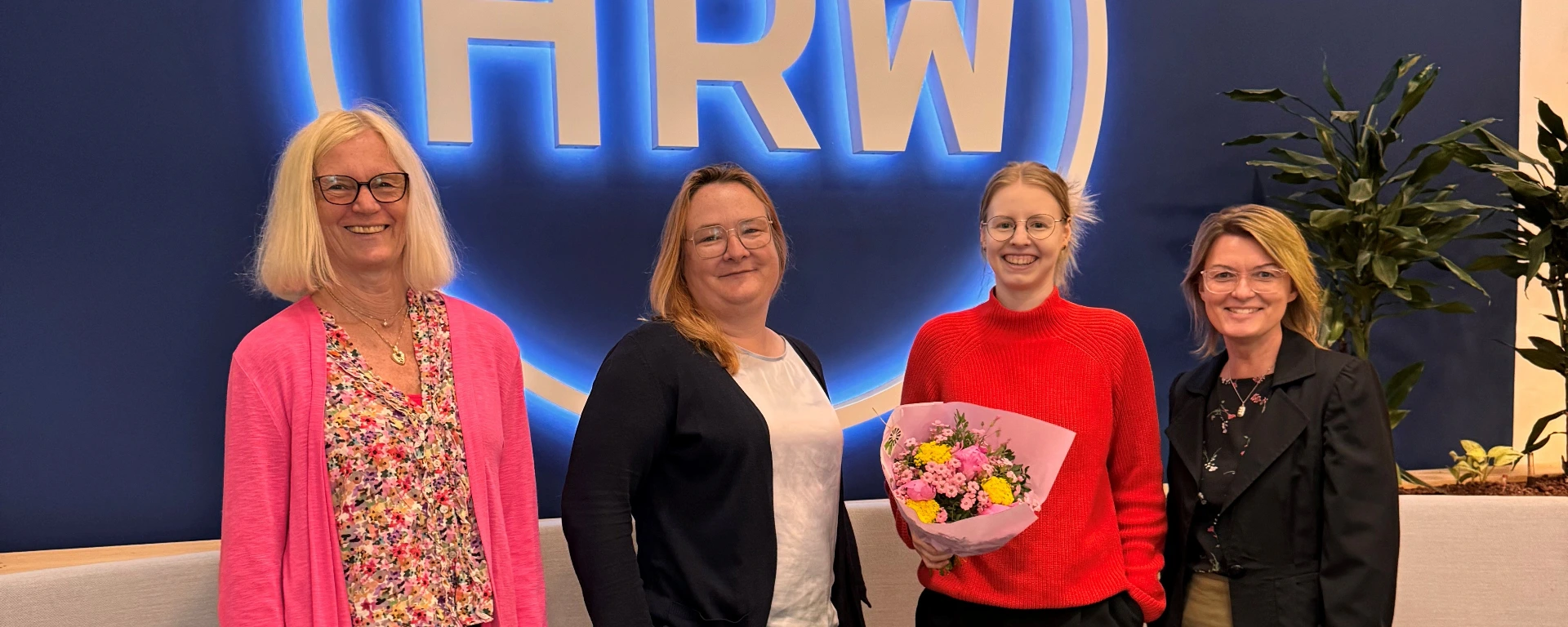 Gruppenfoto an der Hochschule Ruhr West: Antonia Tabea Appel hält einen Blumenstrauß und steht gemeinsam mit Prof. Dr. Simone Roth, Claudia vom Felde und Dr. Hanna Hinrichs vor dem beleuchteten HRW-Logo.