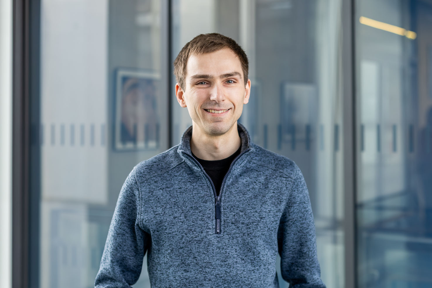 Lächelnde Person in grauem Zip-Pullover vor Glaswänden in moderner Büroumgebung, halbnahes Porträt.