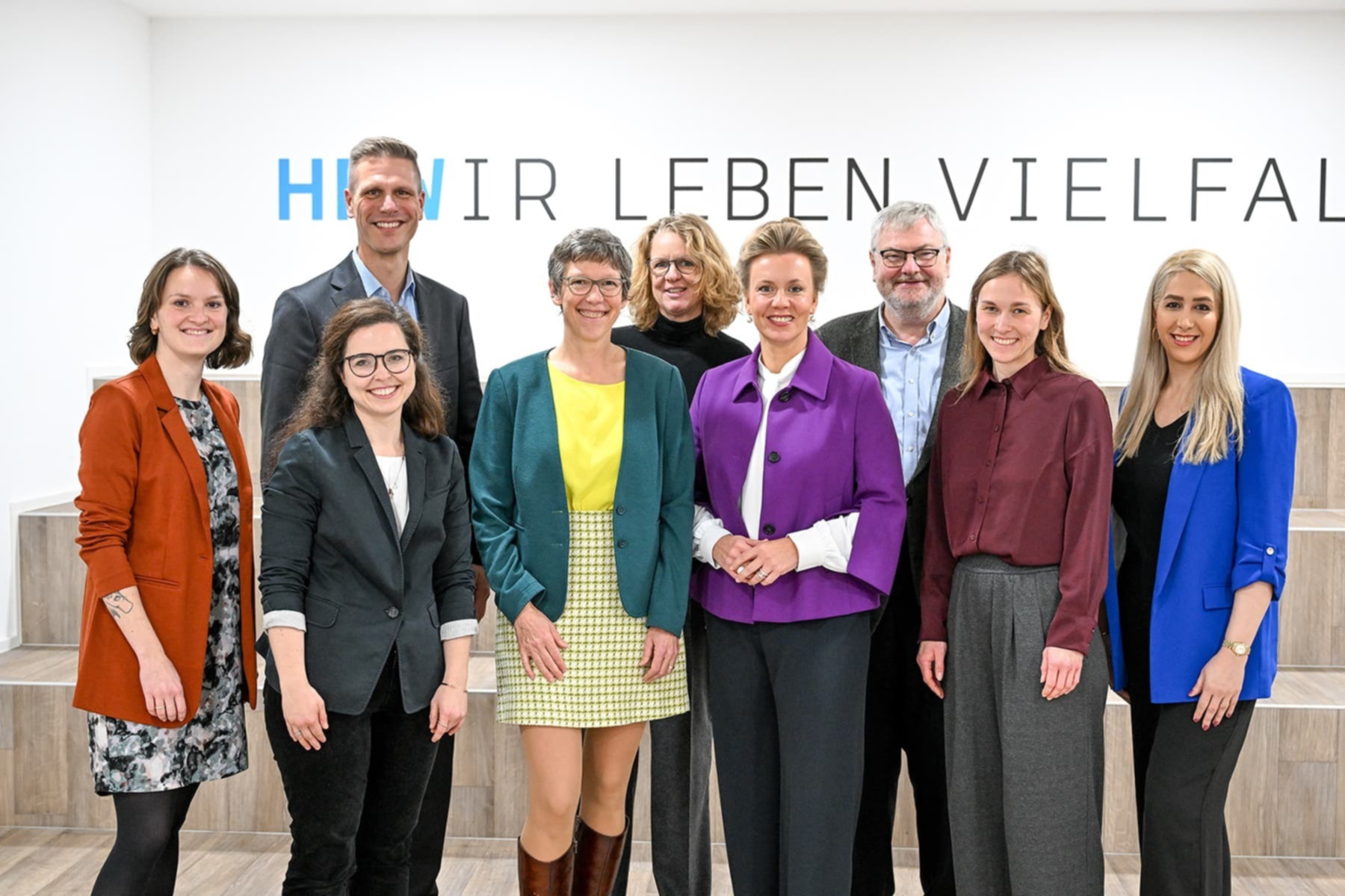 Gruppenfoto von neun Personen, die in einem hellen Raum nebeneinander stehen und in die Kamera lächeln. Im Hintergrund steht der Schriftzug ‚Wir leben Vielfalt‘.