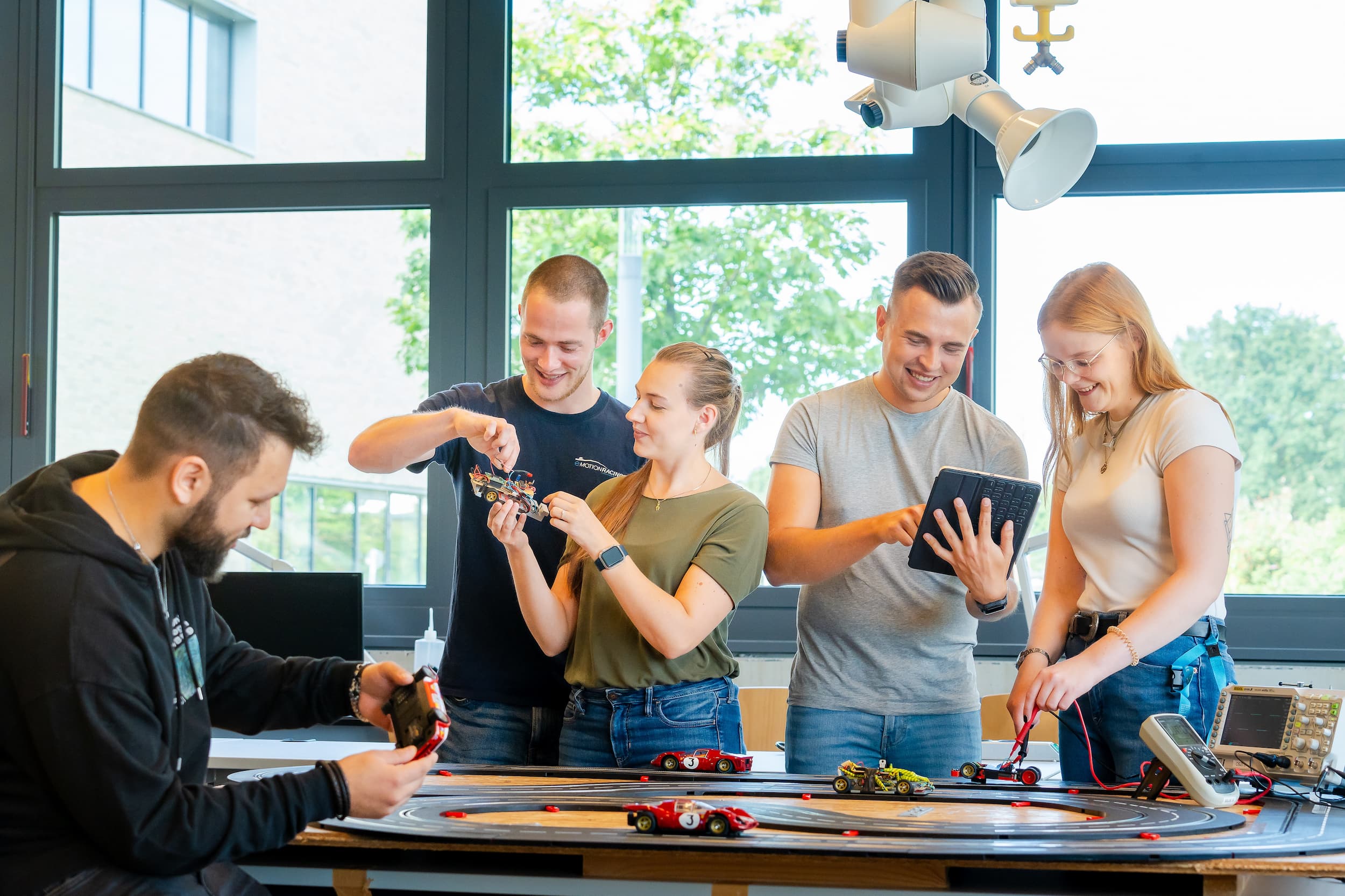 Studierende arbeiten gemeinsam an einer Carrera-Modellrennstrecke; sie prüfen Fahrzeuge, messen Daten und steuern Abläufe per Tablet.
