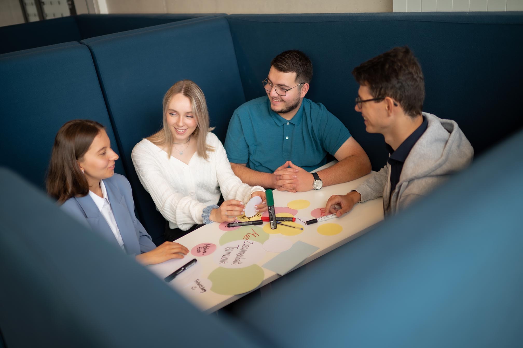 Eine Gruppe von Studierenden sitzt an einem Lernplatz, hält Marker in der Hand, schreibt einige Notizen und lächelt.