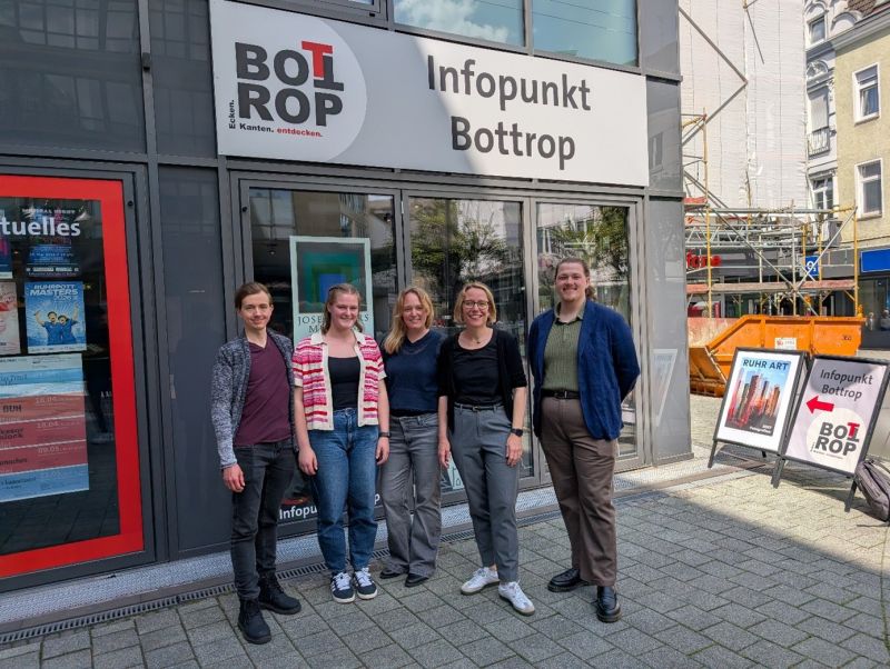 Studierende zusammen mit ihren Betreuer:innen Prof. Sabrina Eimler und Lukas Erle stehen mit einer Vetreterin der Stadt vor dem Infopunkt Bottrop