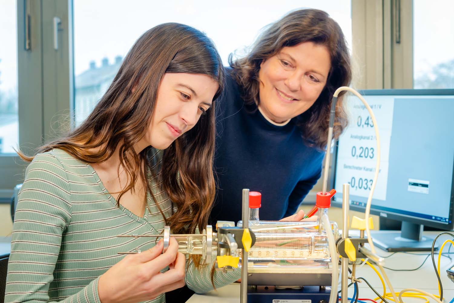 Eine Studierende führt einen physikalischen Versuch durch. Eine Lehrende schaut wohlwollend lächelnd zu.