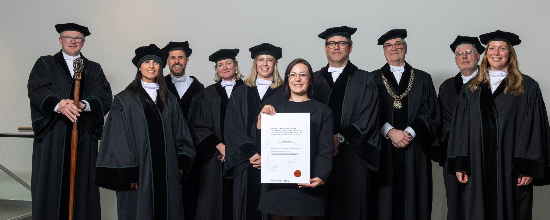 Maike Hübner steht in der Mitte eines Gruppenfotos und hält ihre Promotionsurkunde in die Kamera. Um sie herum stehen weitere Personen in schwarzer akademischer Festkleidung mit Baretts auf einer Bühne vor hellem Hintergrund. Einige tragen repräsentative Insignien.