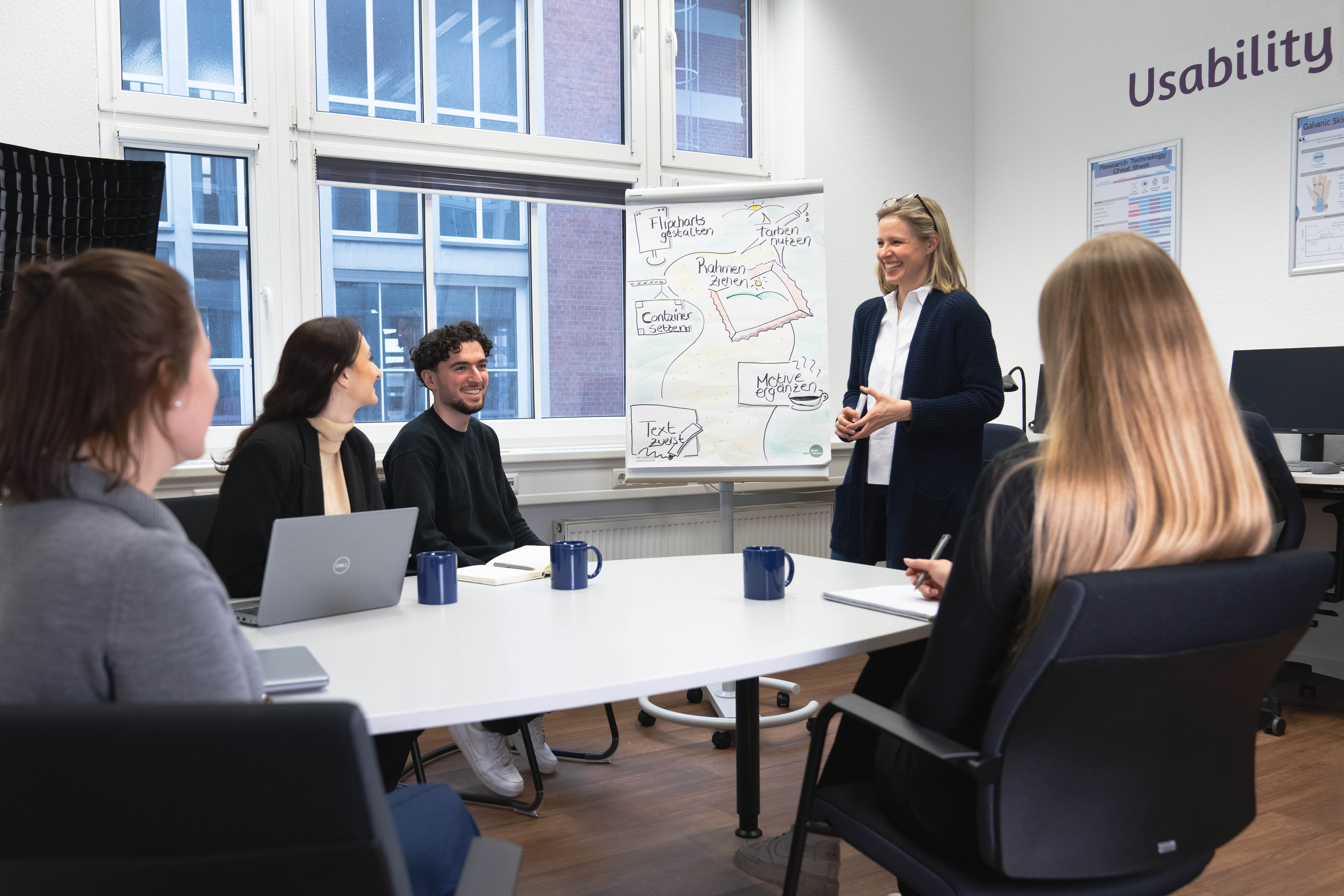 Das Foto zeigt eine Gruppe von Personen, die an einem Besprechungstisch sitzen. Eine Person steht an einem Whiteboard.