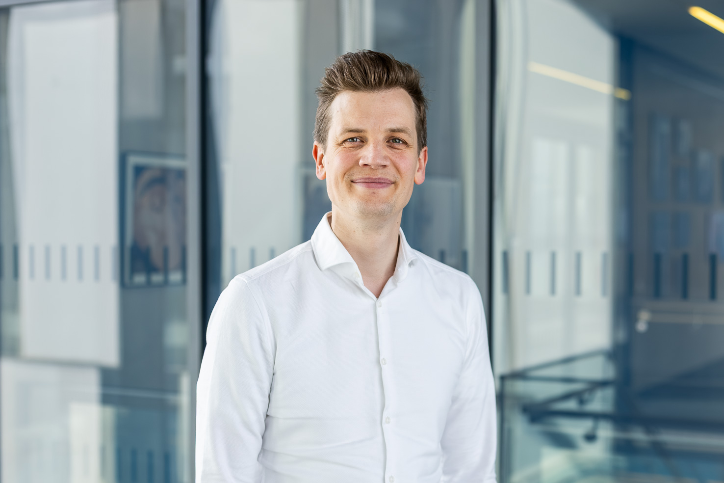 Lächelnder Mann im weißen Hemd in einem modernen Büroflur mit Glaswänden.