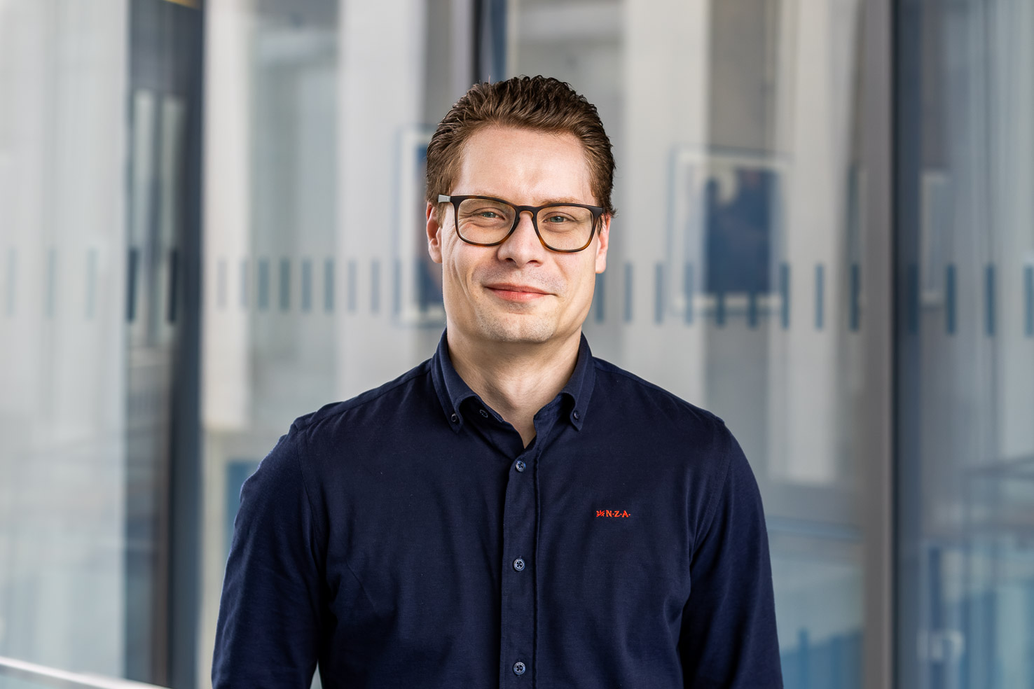 Porträt eines Mannes mit Brille, dunkelblaues Hemd, lächelt vor verglaster Bürowand mit Dekorstreifen.