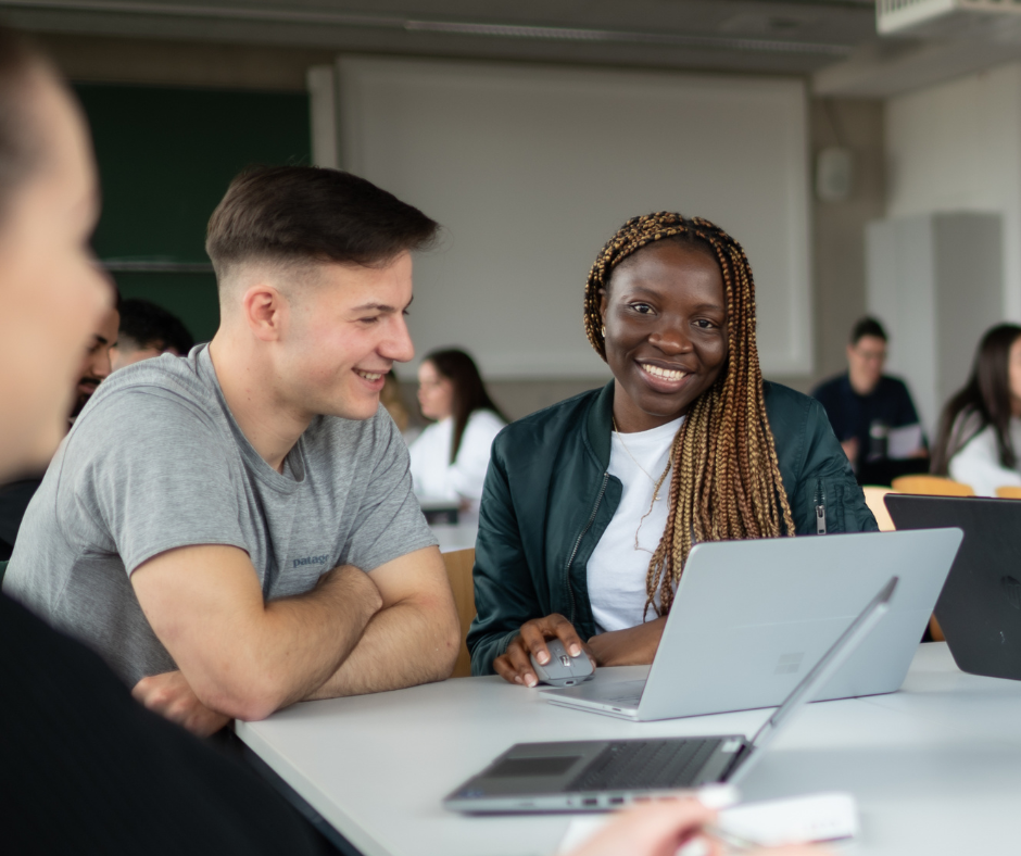 Studierende in einem Seminarraum. Ein Student und eine Studierende vor einem Laptop. Sie lächelt in die Kamera