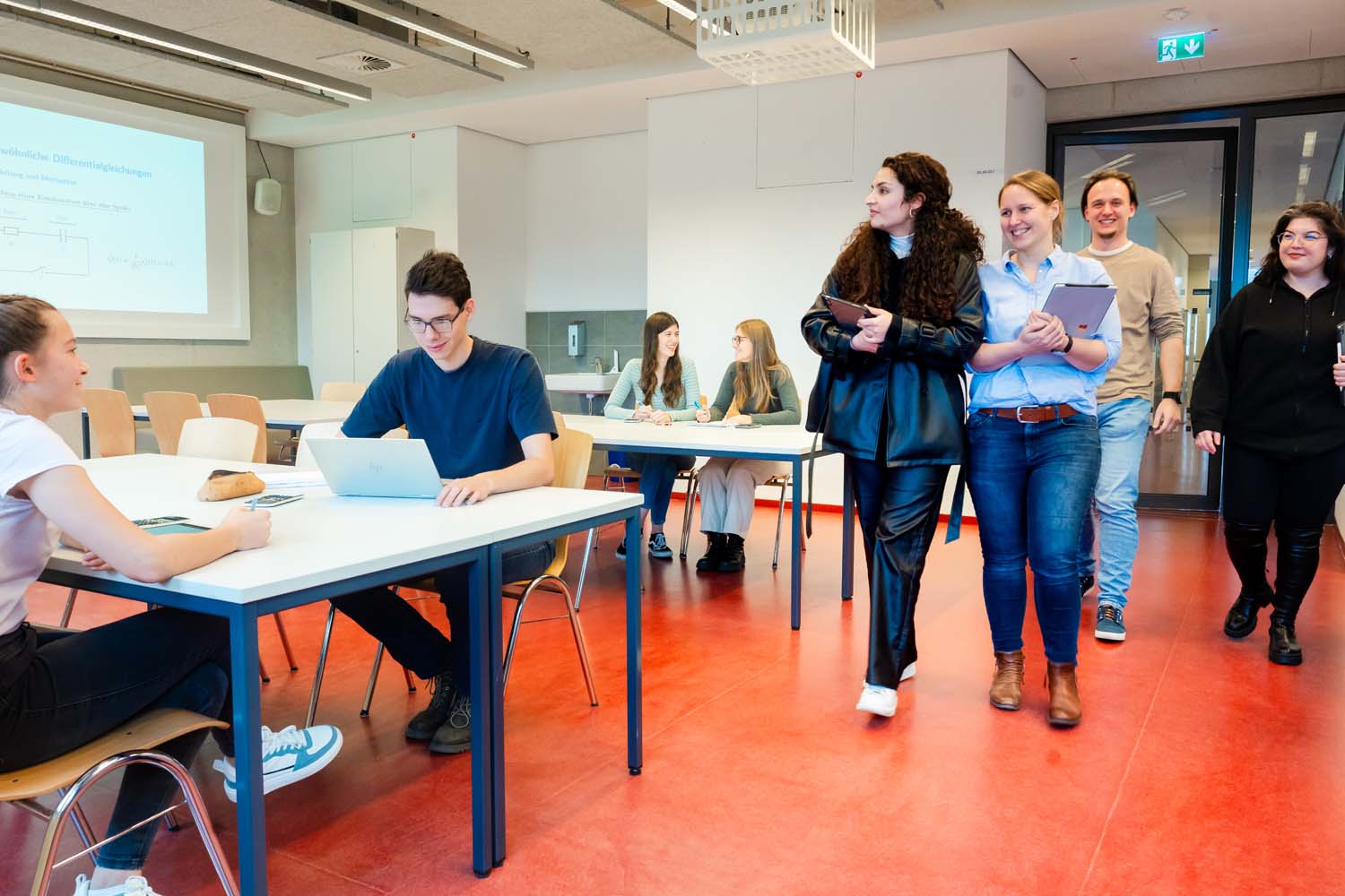 Gäste betreten einen Lernraum, in dem bereits Studierende an Tischen sitzen. Im Hintergrund ist eine Folie an die Wand projiziert.