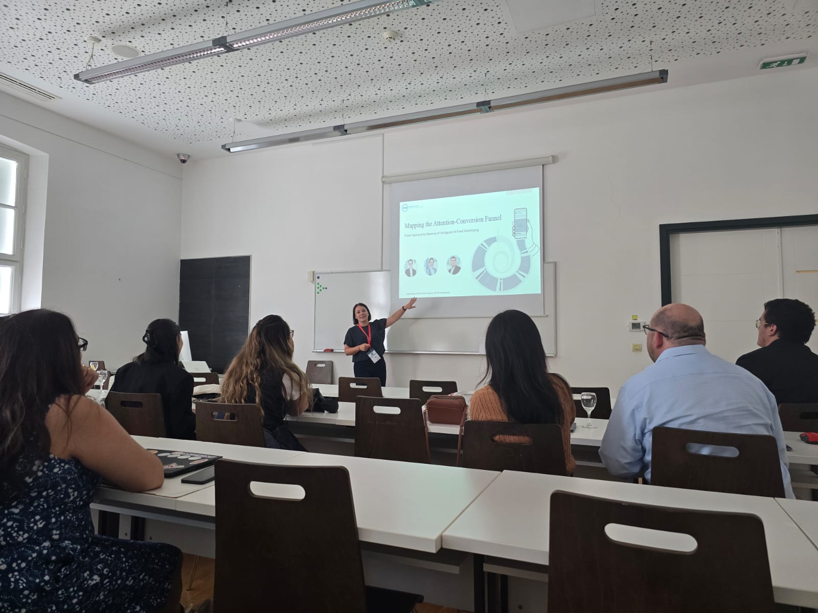 Maike Hübner hält einen Vortrag in einem Seminarraum und zeigt auf eine Präsentationsfolie mit dem Titel „Mapping the Attention-Conversion Funnel“. Mehrere Personen sitzen an Tischen und hören zu.