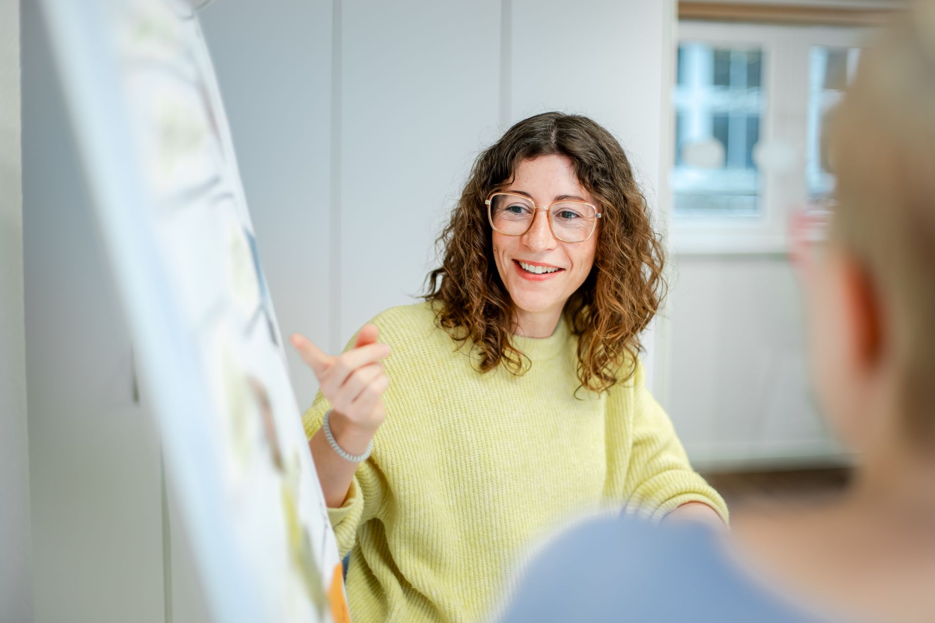 Eine Frau mit lockigem Haar und Brille erklärt lächelnd etwas an einem Flipchart. Die Szene wirkt wie eine kreative Besprechung oder ein Workshop in freundlicher Atmosphäre