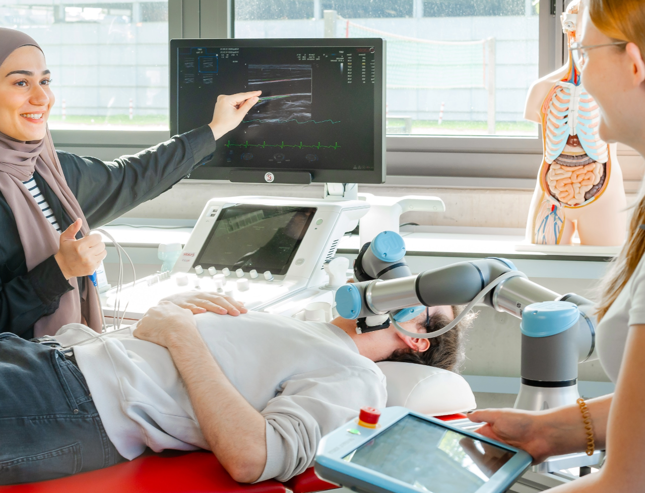 Zwei weibliche Studierende arbeiten an einem Ultraschallgerät. Zwischen ihnen liegt ein männlicher Proband mit einer Atemmaske