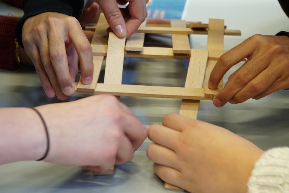 das Bild zeigt vier Hände, die gemeinsam eine Leonardo-Brücke aus Holz bauen
