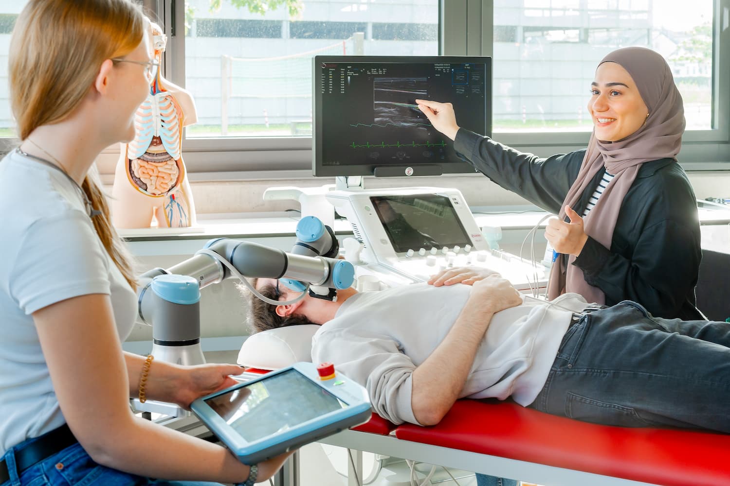 Studierende führen im Labor eine Ultraschalluntersuchung mit einem robotischen Assistenzarm durch; eine Person liegt auf der Untersuchungsliege, eine andere zeigt auf das Ultraschallbild.
