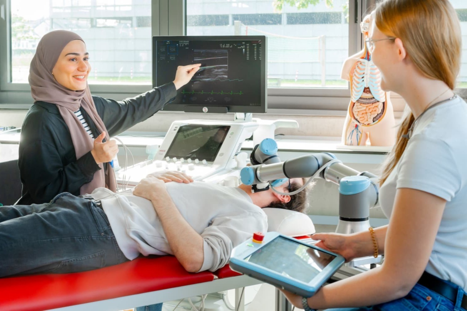Studierende führen im Labor eine Ultraschalluntersuchung mit einem robotischen Assistenzarm durch; eine Person liegt auf der Untersuchungsliege, eine andere zeigt auf das Ultraschallbild.
