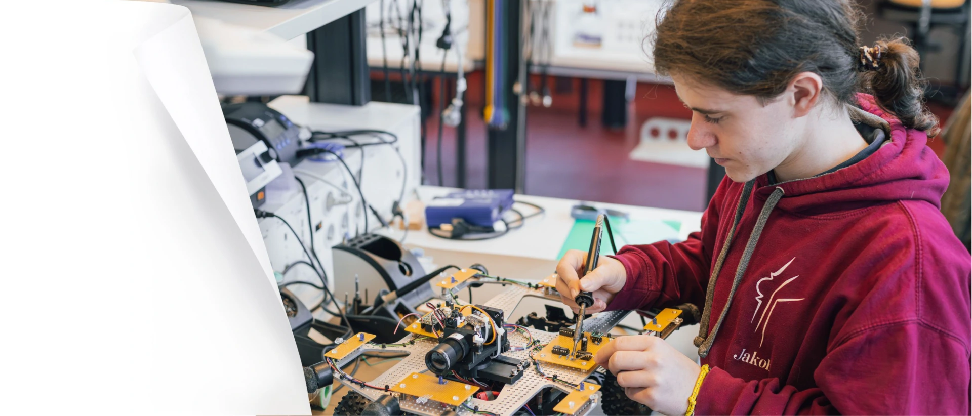 Person im roten Hoodie arbeitet an einem offenen Roboter-/Elektronikaufbau auf einer Werkbank und verschraubt Bauteile mit einem Schraubendreher; im Hintergrund Laborgeräte und Kabel.