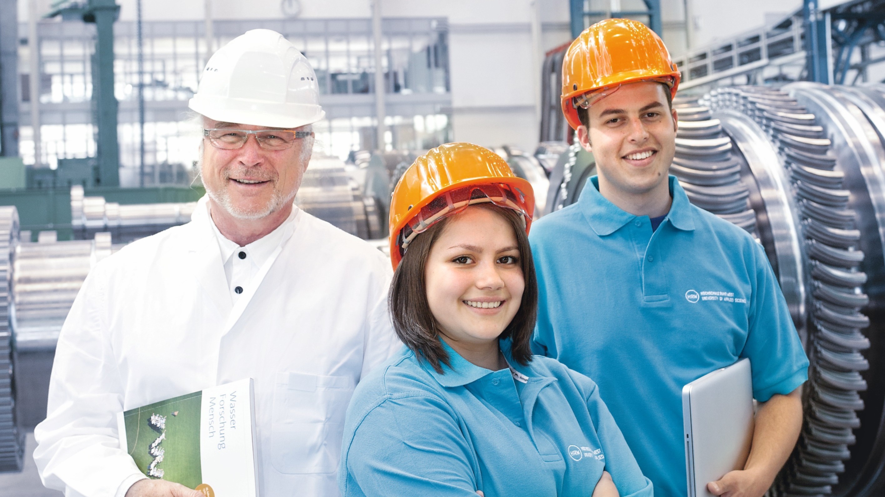 Zwei Studierende und ein Unternehmer mit Helmen in einer Werkstatt