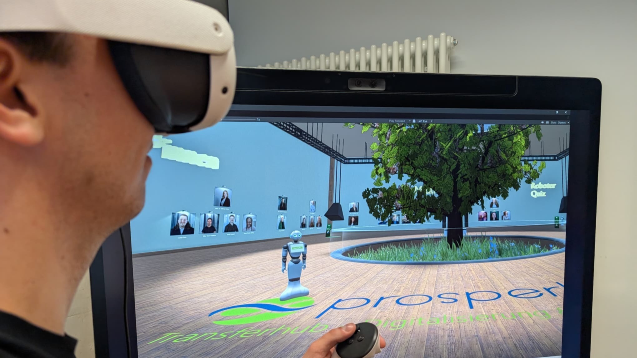 Eine Person trägt eine Virtual-Reality-Brille und hält einen Controller in der Hand. Auf einem großen Bildschirm ist eine virtuelle Umgebung mit einem Baum, einem kleinen Roboter und eingeblendeten Profilbildern zu sehen.