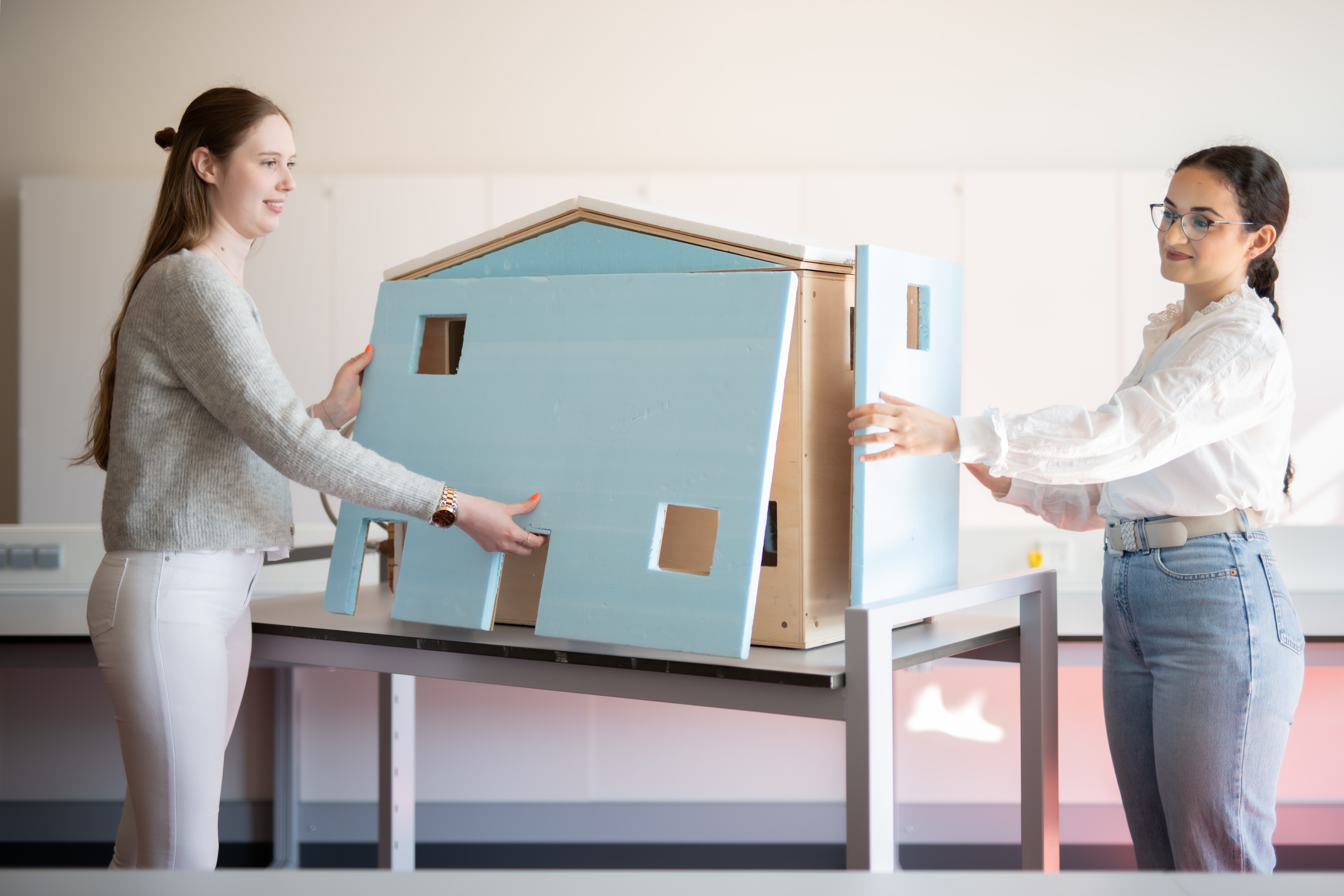 Das Foto zeigt zwei Studentinnen, die an einem Modellhaus stehen und jeweils ein Dämmmaterial in der Hand halten und es an das Modellhaus anbringen.