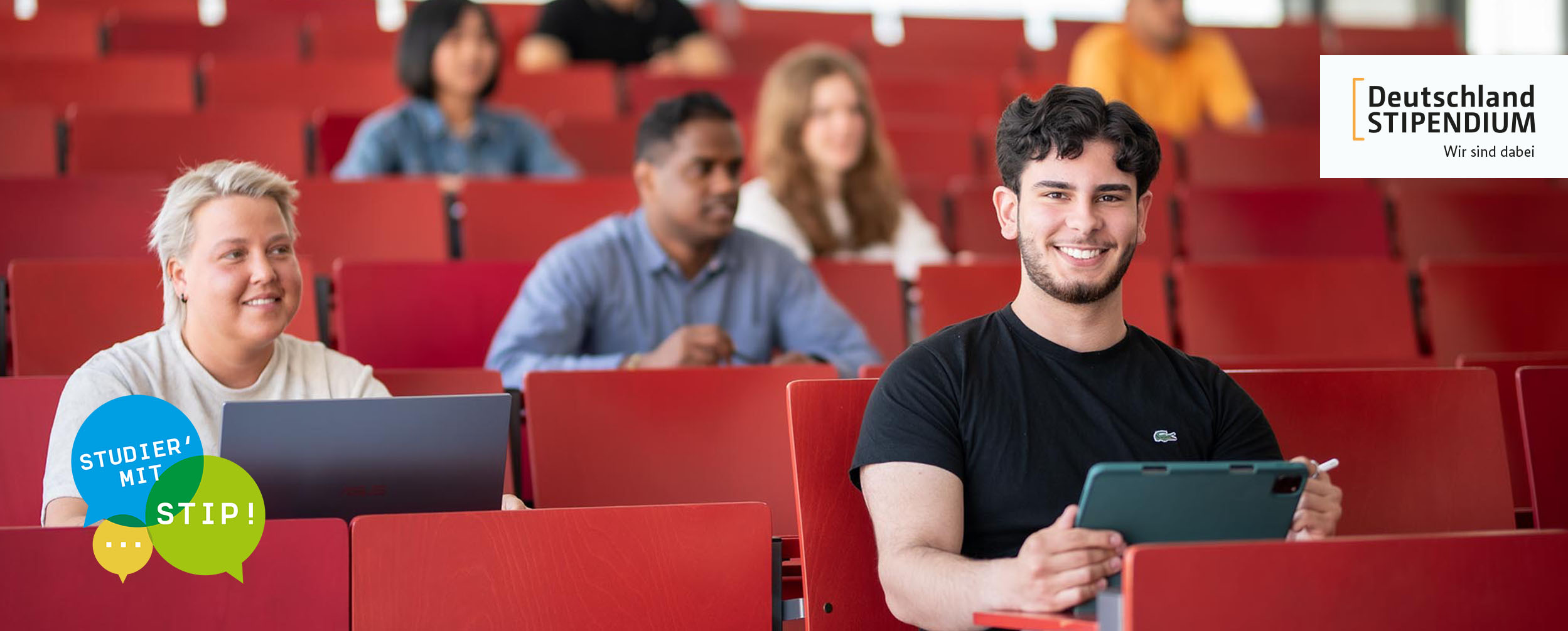 Studierende sitzen im Hörsaal. Im Fokus ist ein junger Mann mit Tablet