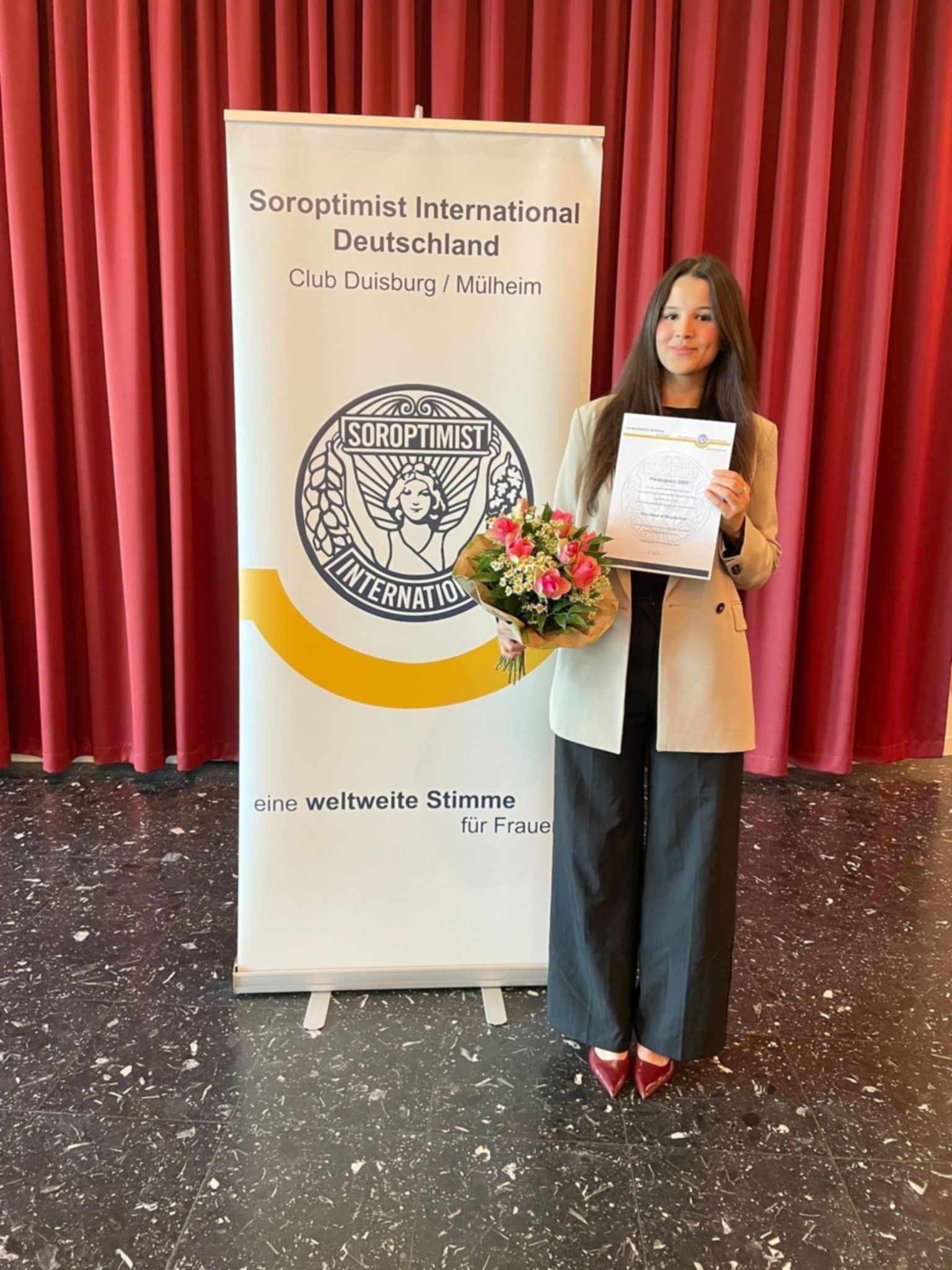 Eine Studentin hält ihre Urkunde und steht vor einem Aufsteller