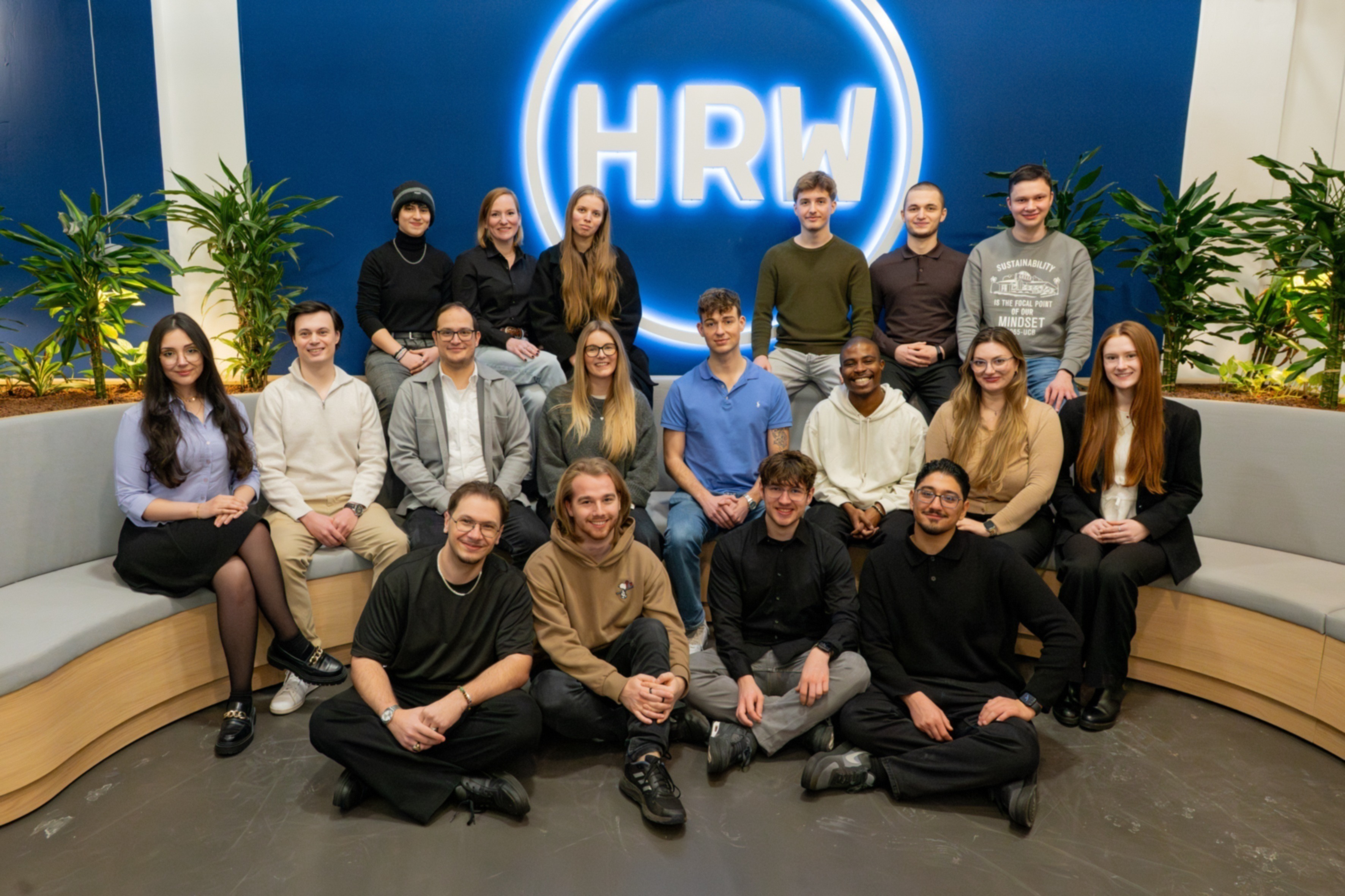 Frauen und Männer sitzen auf und vor einer Bank, einige Personen stehen dahinter