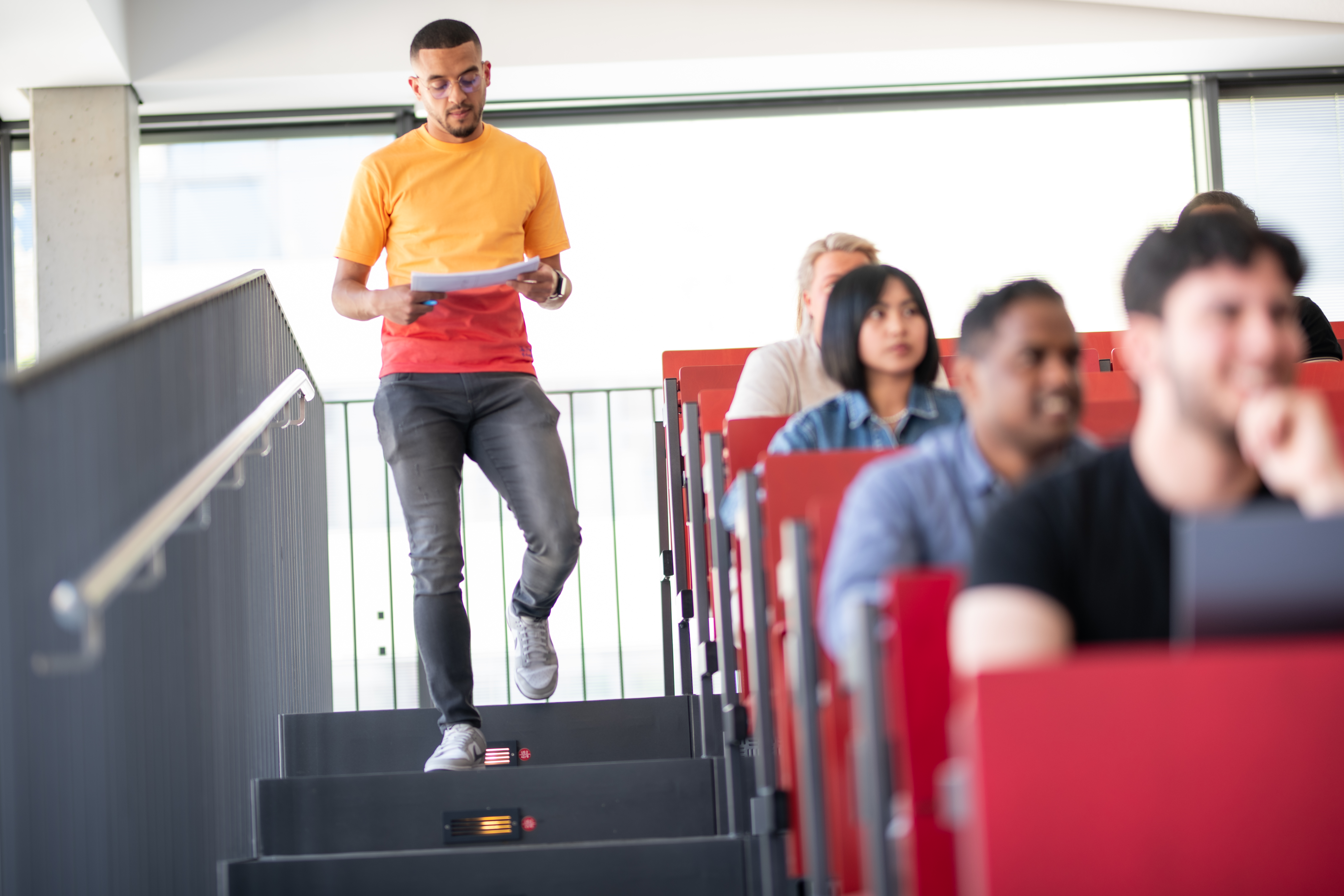 Das Foto zeigt einen Hörsaal mit diversem Publikum. Im Hintergrund läuft ein Student die Treppe runter.