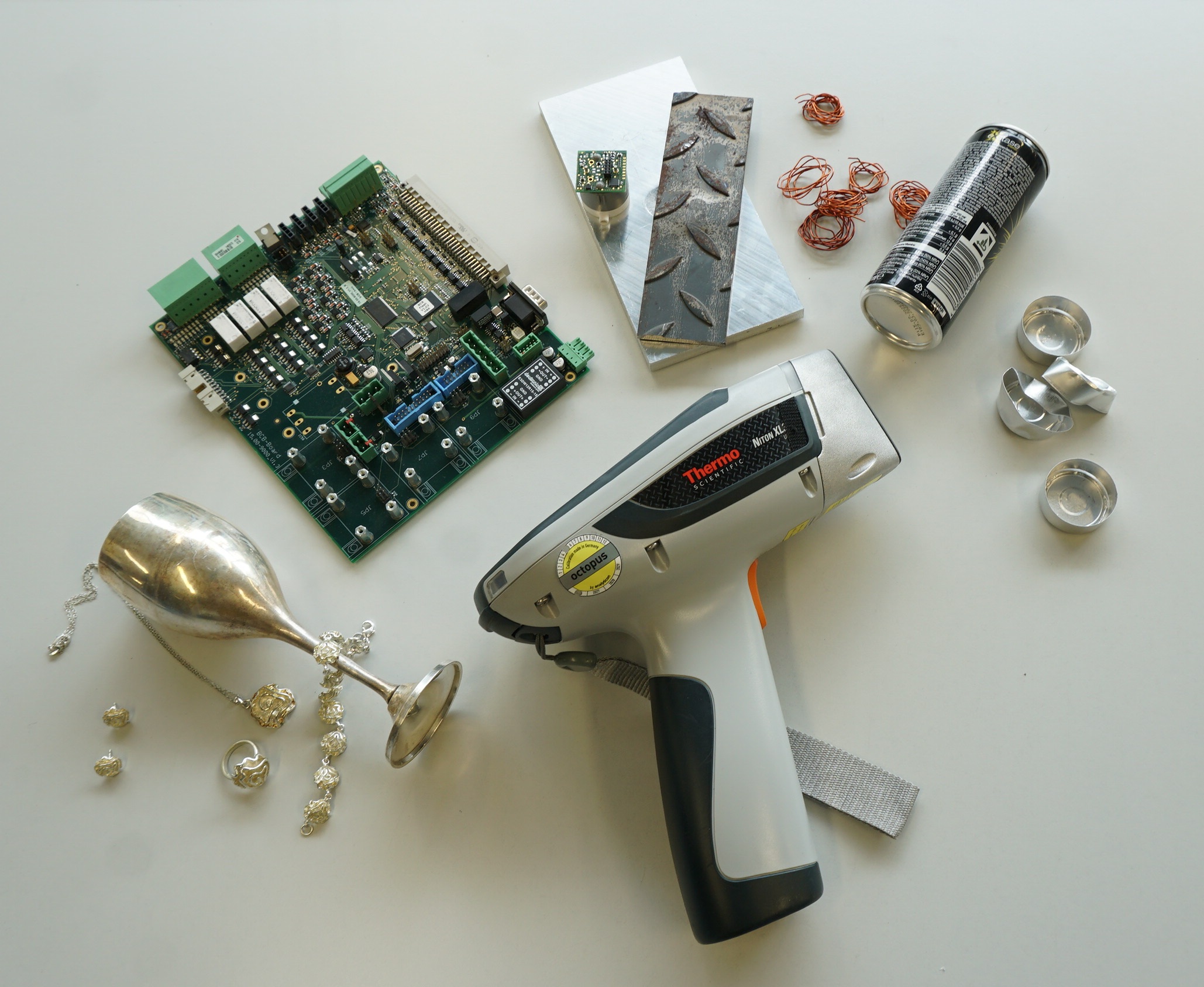 Auf einem Tisch: tragbarer Metallanalysator, Leiterplatte, Metallreste, Kupferdraht, Aluminiumteile, umgestürzter Kelch und kleine Dose.