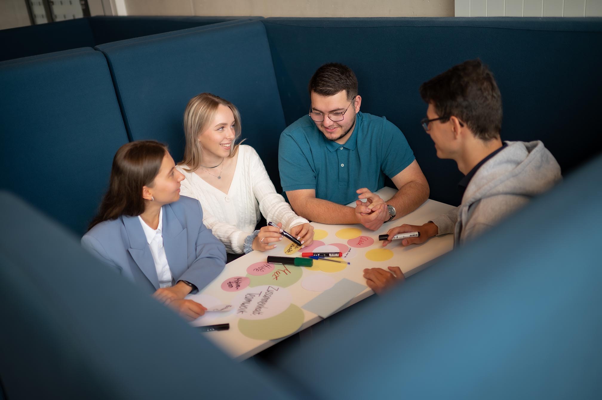 Eine Gruppe von Studierenden sitzt an einem Lernplatz, hält Marker in der Hand, schreibt einige Notizen und lächelt.